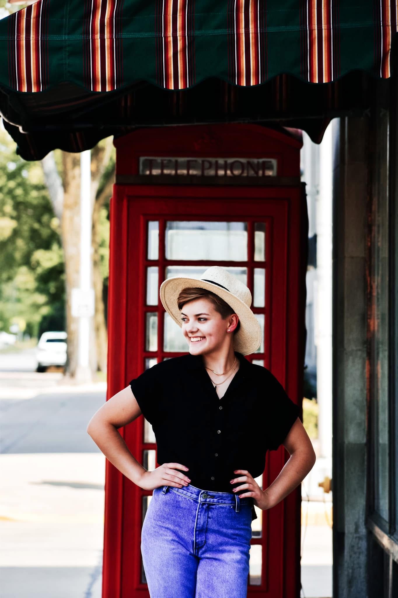 A woman in a hat is standing in front of a red telephone booth.
