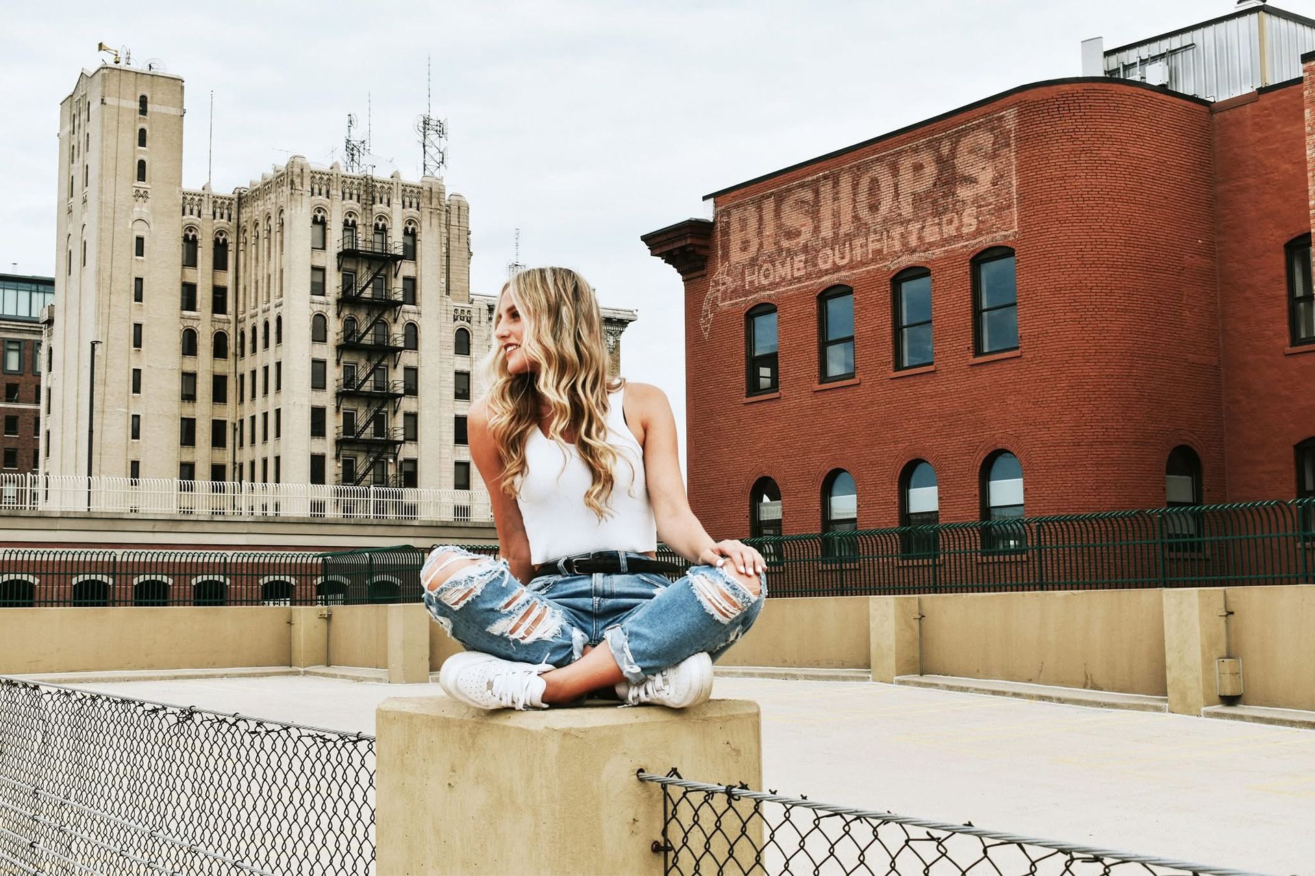 A woman is sitting on a concrete block with her legs crossed in front of a brick building.