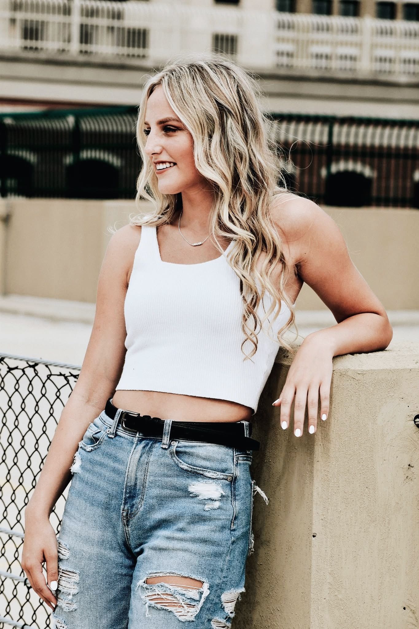 A woman in a white tank top and ripped jeans is leaning against a wall.