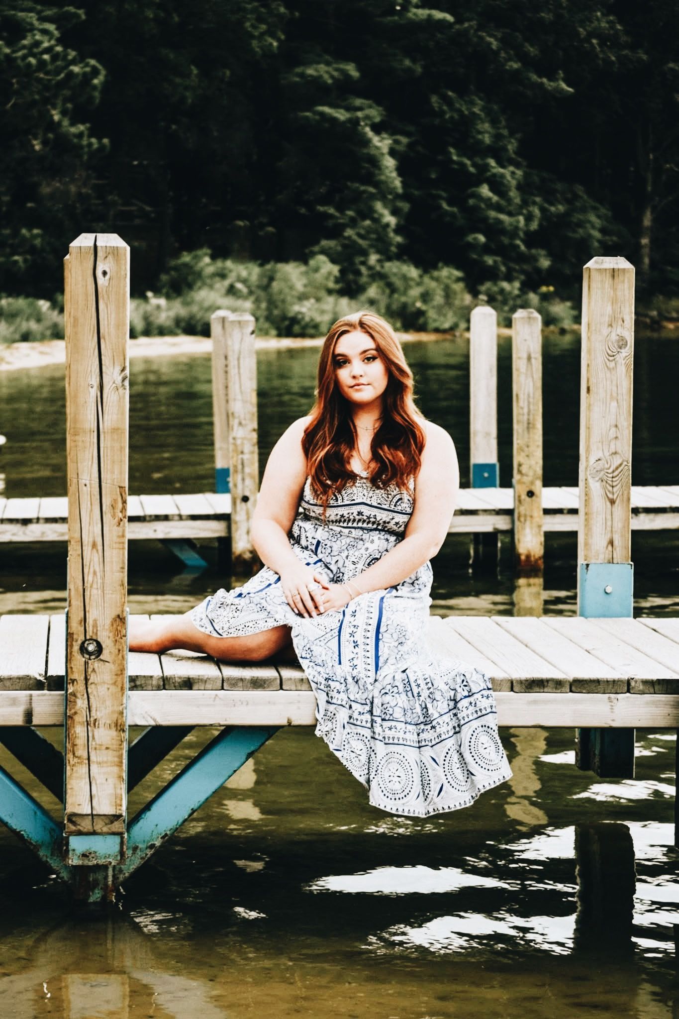 A woman in a dress sits on a dock overlooking a body of water