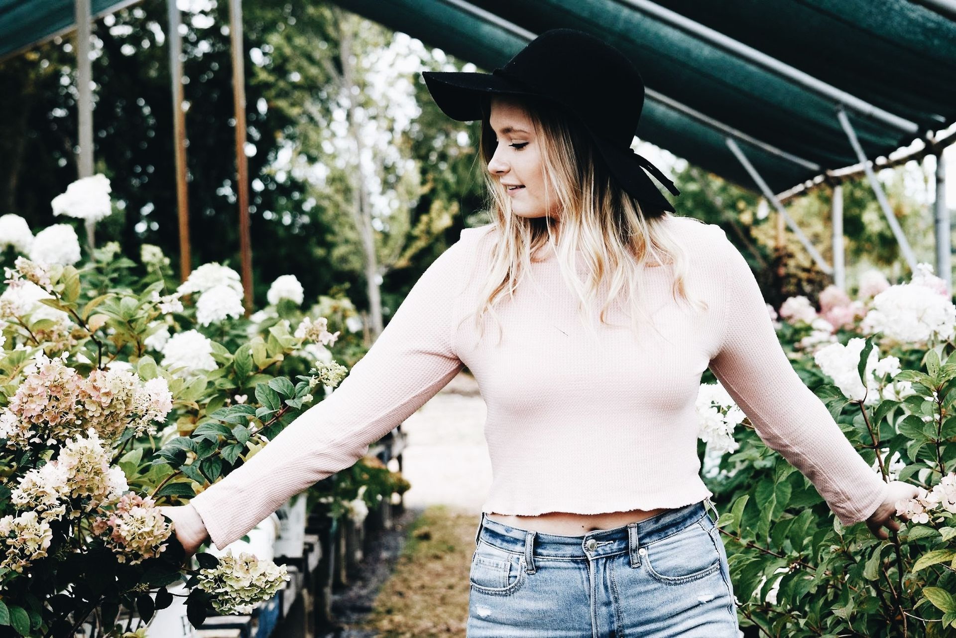 A woman in a hat is standing in a greenhouse with her arms outstretched.