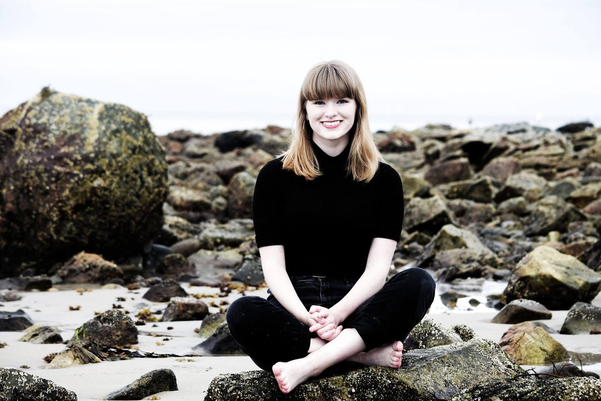 A woman is sitting on a rock on the beach with her legs crossed.