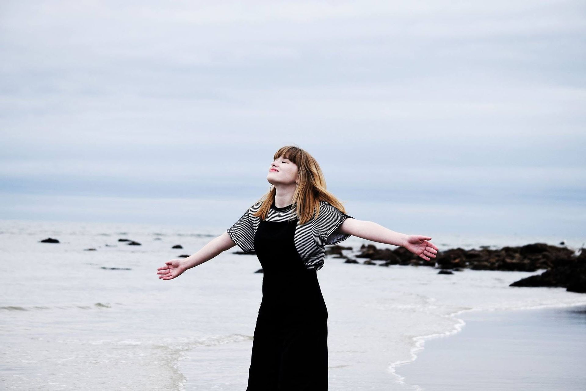 A woman in a black dress is standing on a beach with her arms outstretched.