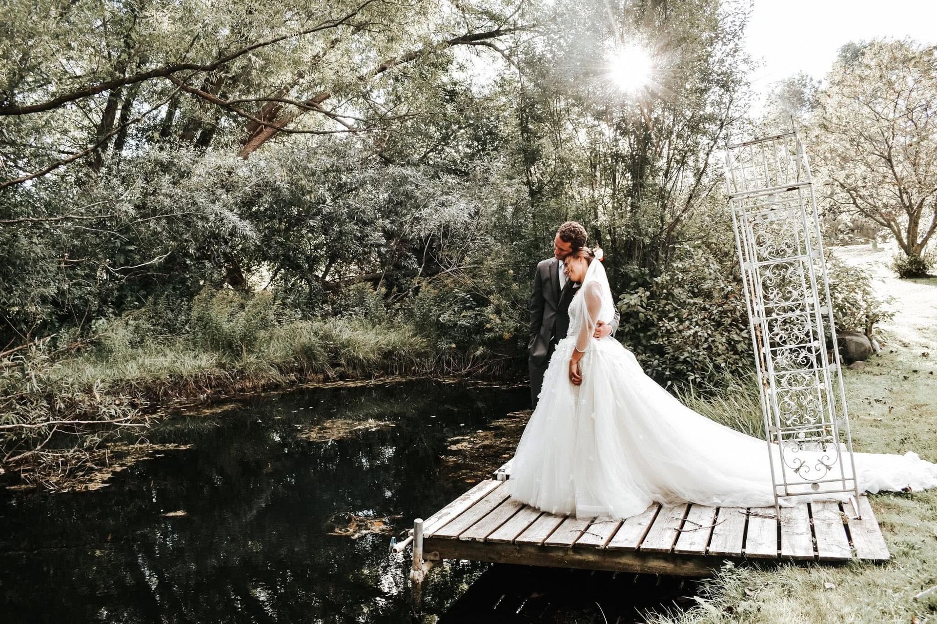 A bride and groom are standing on a wooden dock next to a pond.