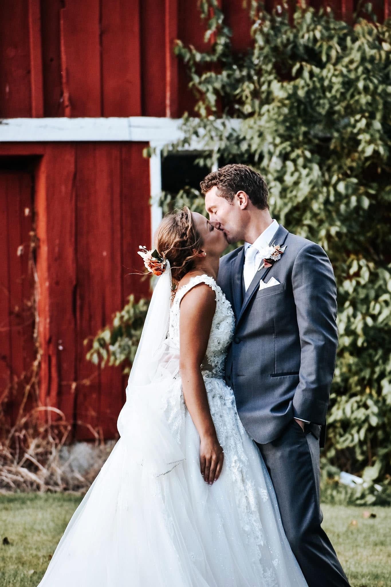 A bride and groom are kissing in front of a red barn.