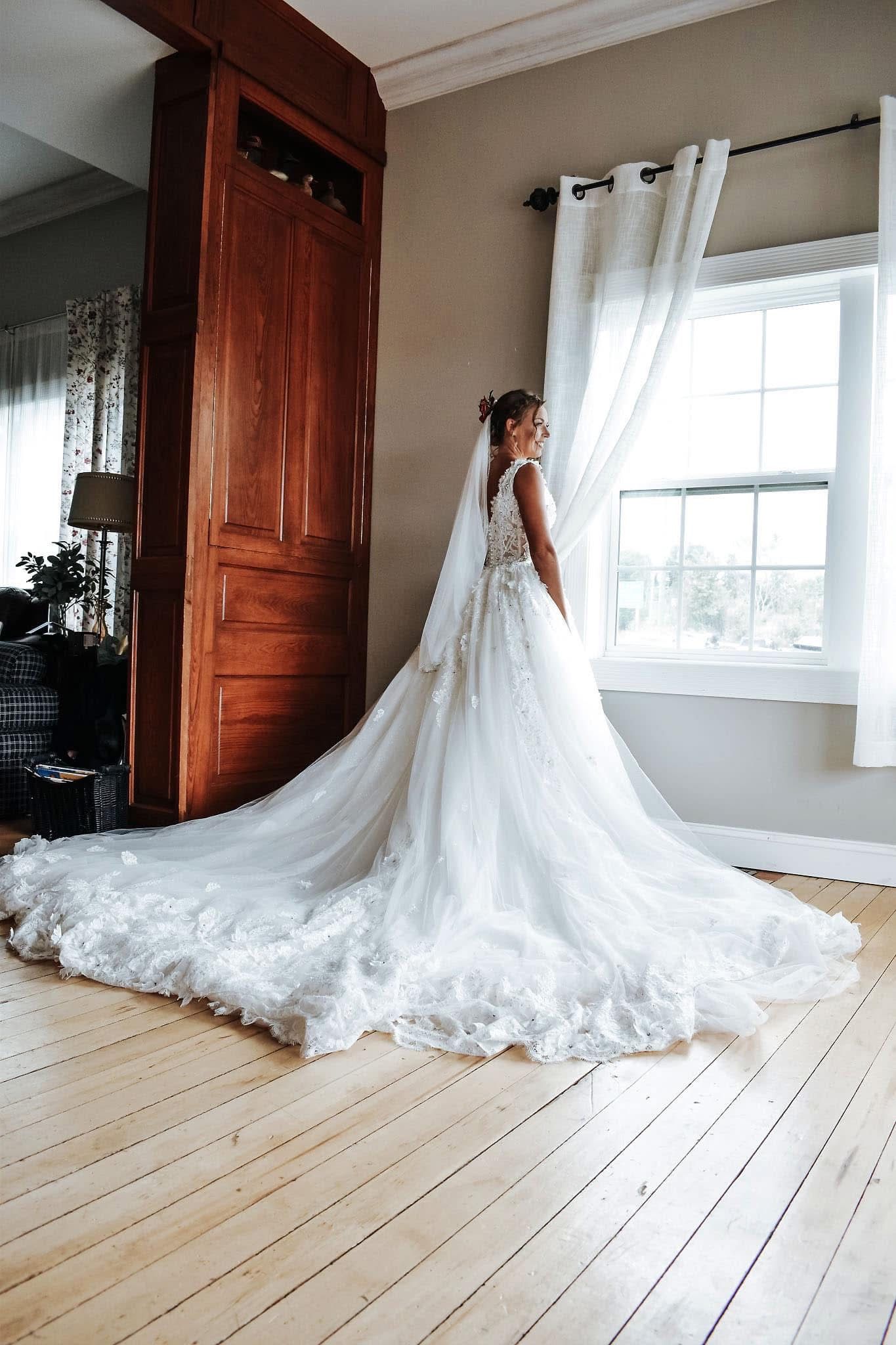 A bride in a long wedding dress is standing in front of a window.