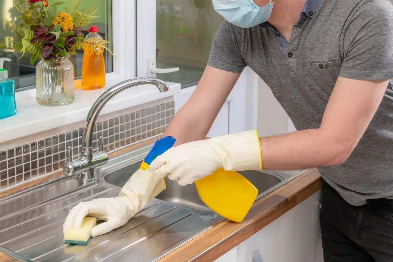 A man wearing a mask and gloves is cleaning a kitchen sink.