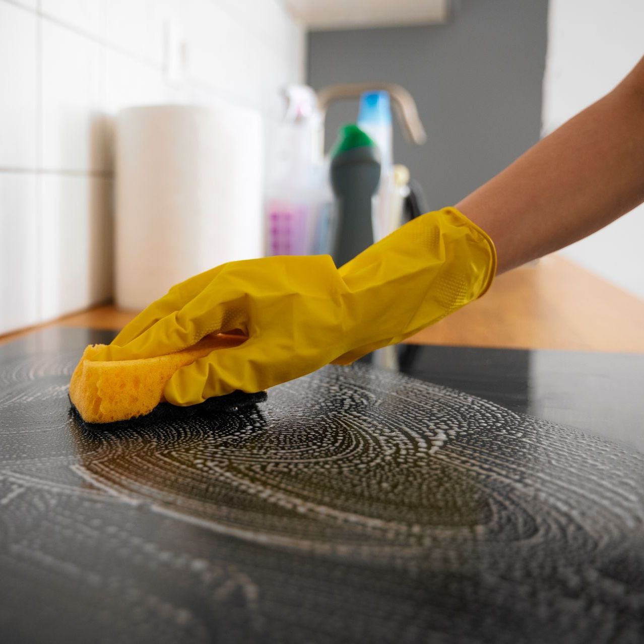 A person wearing yellow gloves is cleaning a stove top with a sponge.