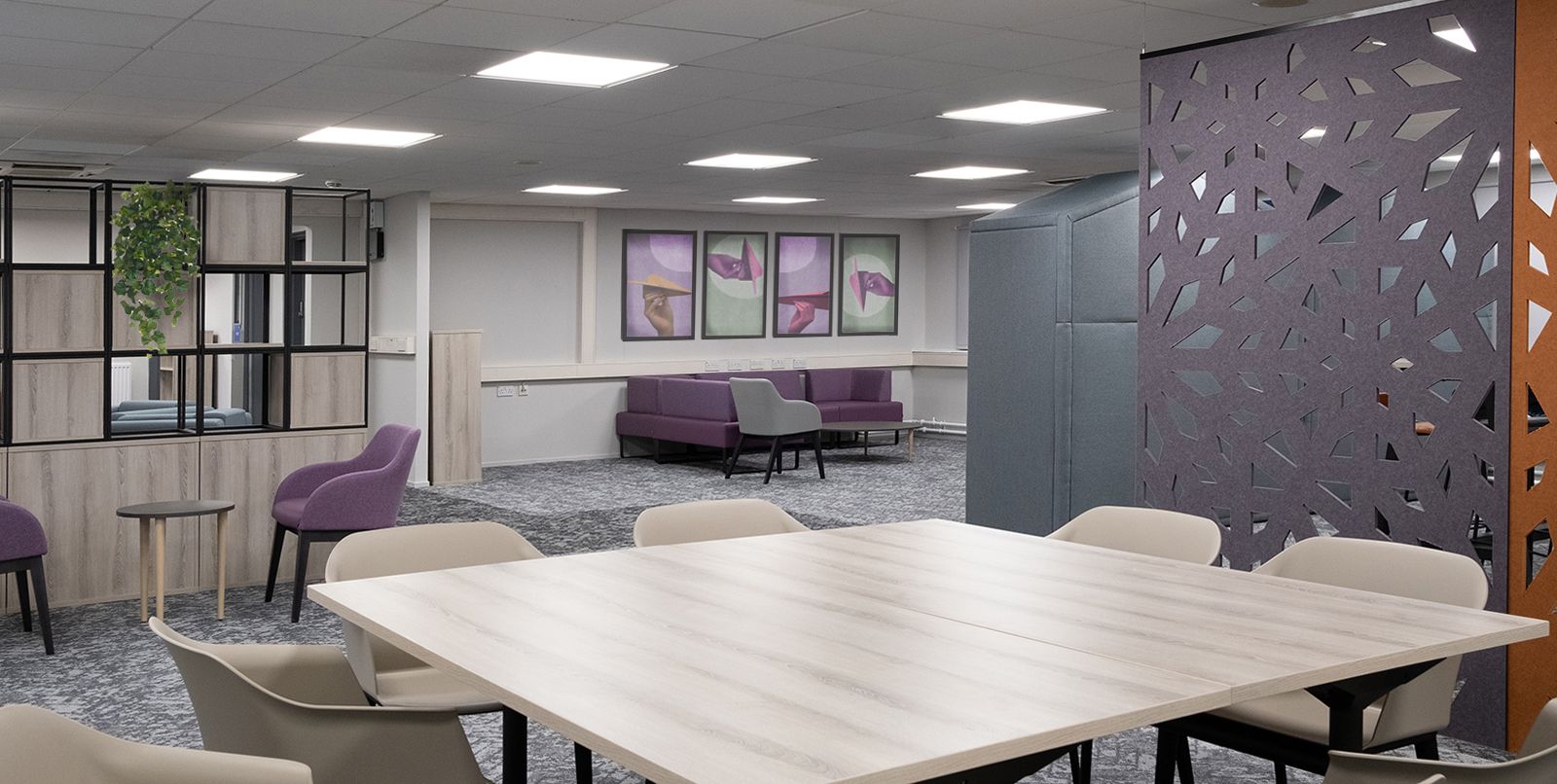 At the forefront of the image is a large wooden desk with 8 chairs around it. In the back of the room is a purple sofa underneath four large art pieces on the wall. The colours of the room are grey, purple, and green.