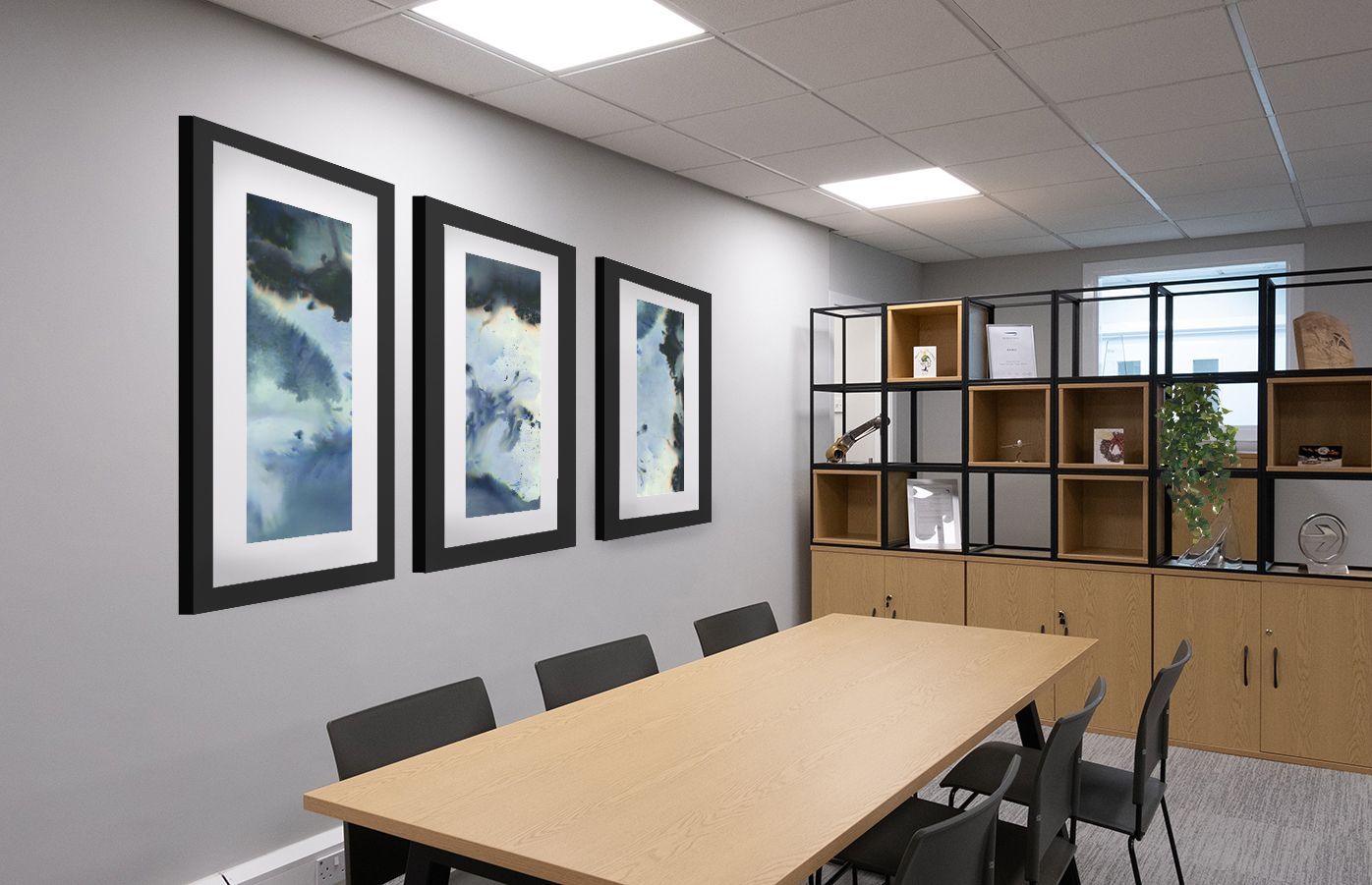 There is a wooden table with six black chairs. On the wall behind the table are three black-framed photos. There is a storage divider at the end of the table, showcasing art, credentials, and plants.