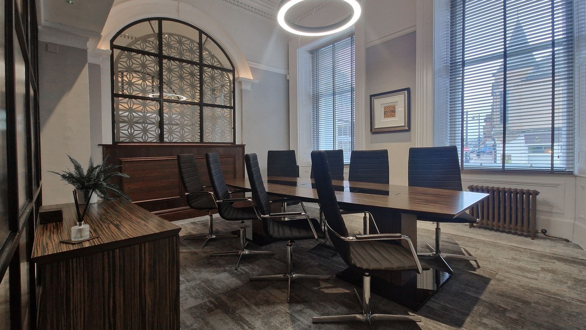Meeting room at William Duncan office. There is a dark wood table in the middle of the room with eight task chairs. There are two large windows behind the desk and a circular overhead light.