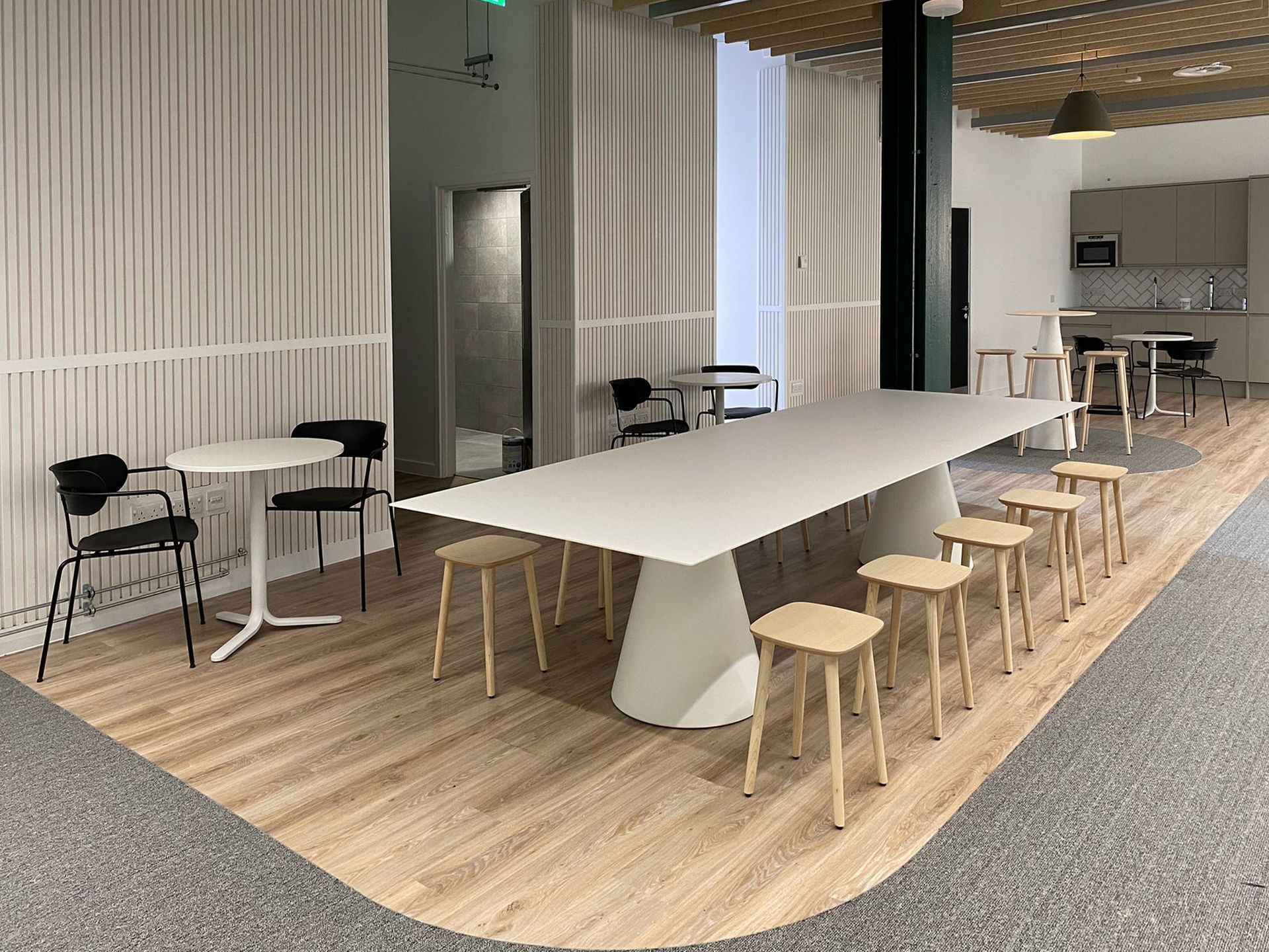 A kitchen and dining area with a large white conference table and oak stools. There is a mixture of low and high circular tables with dining chairs and tall stools. The kitchen area has greige cabinets, a sink, microwave, and fridge section, all integrated into the space. The walls and ceiling have acoustic panelling to help with soundproofing.