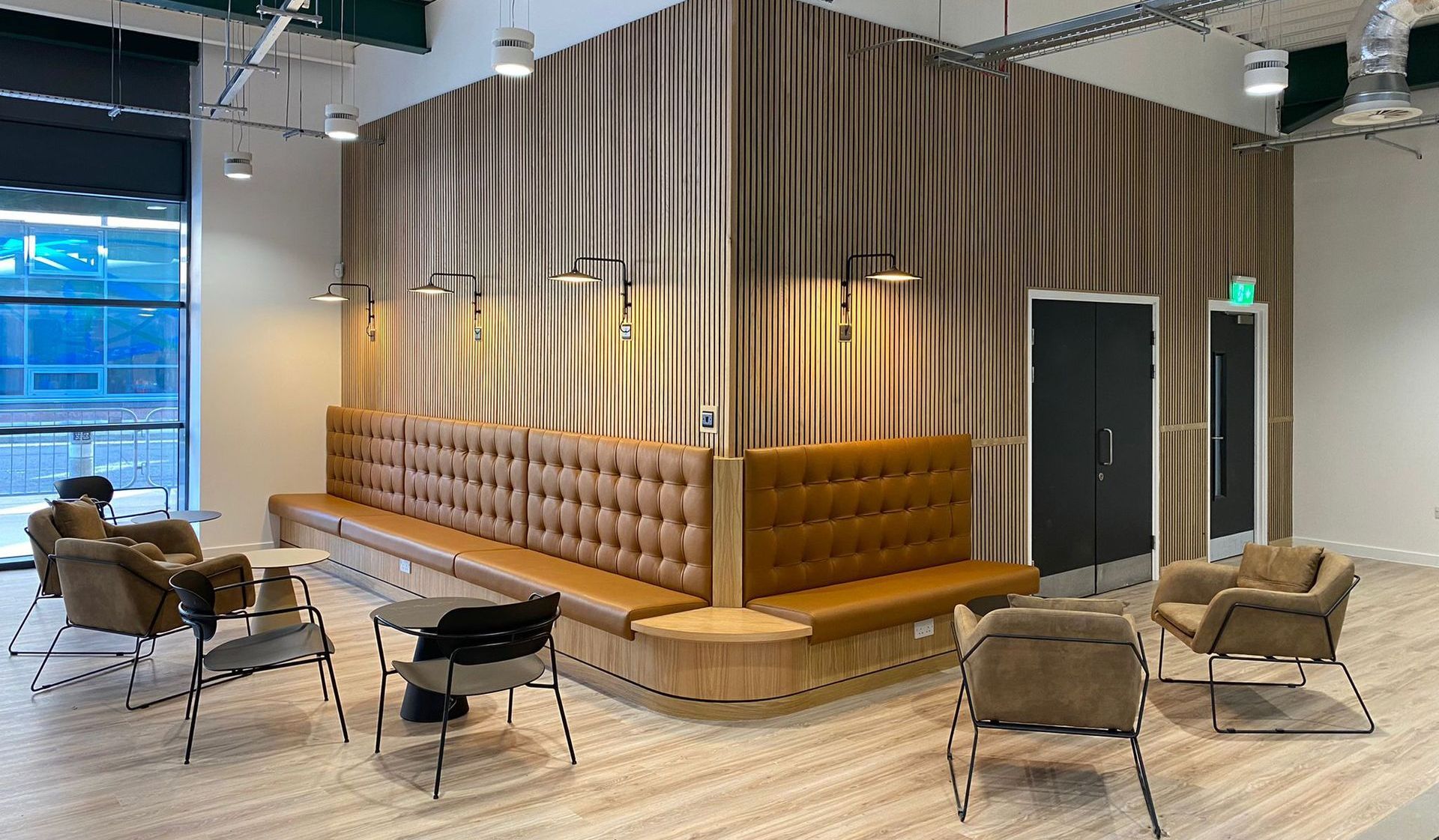 Open area near large windows with a corner wall featuring thin wooden panelling. Against the wall are tan leather integrated benching seating with plug sockets beneath. There are three circular coffee tables placed nearby with a mixture of upholstered and non-upholstered armchairs, to create a collaborative casual area. 