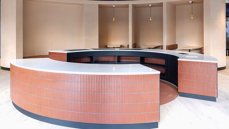 A large circular reception table in an open plan building. The tile is a pinky orange colour with blue detailing and white countertops. In the background are three seating booths with tables and hanging lights. 