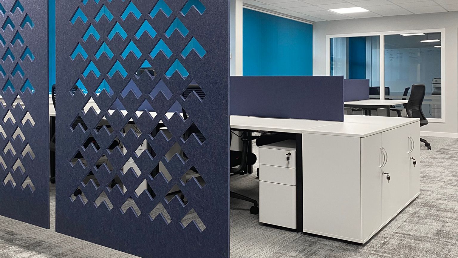 A staff lounge/office space with white desks and black office chairs. The room is punctuated with blue acoustic panels and dividers. 