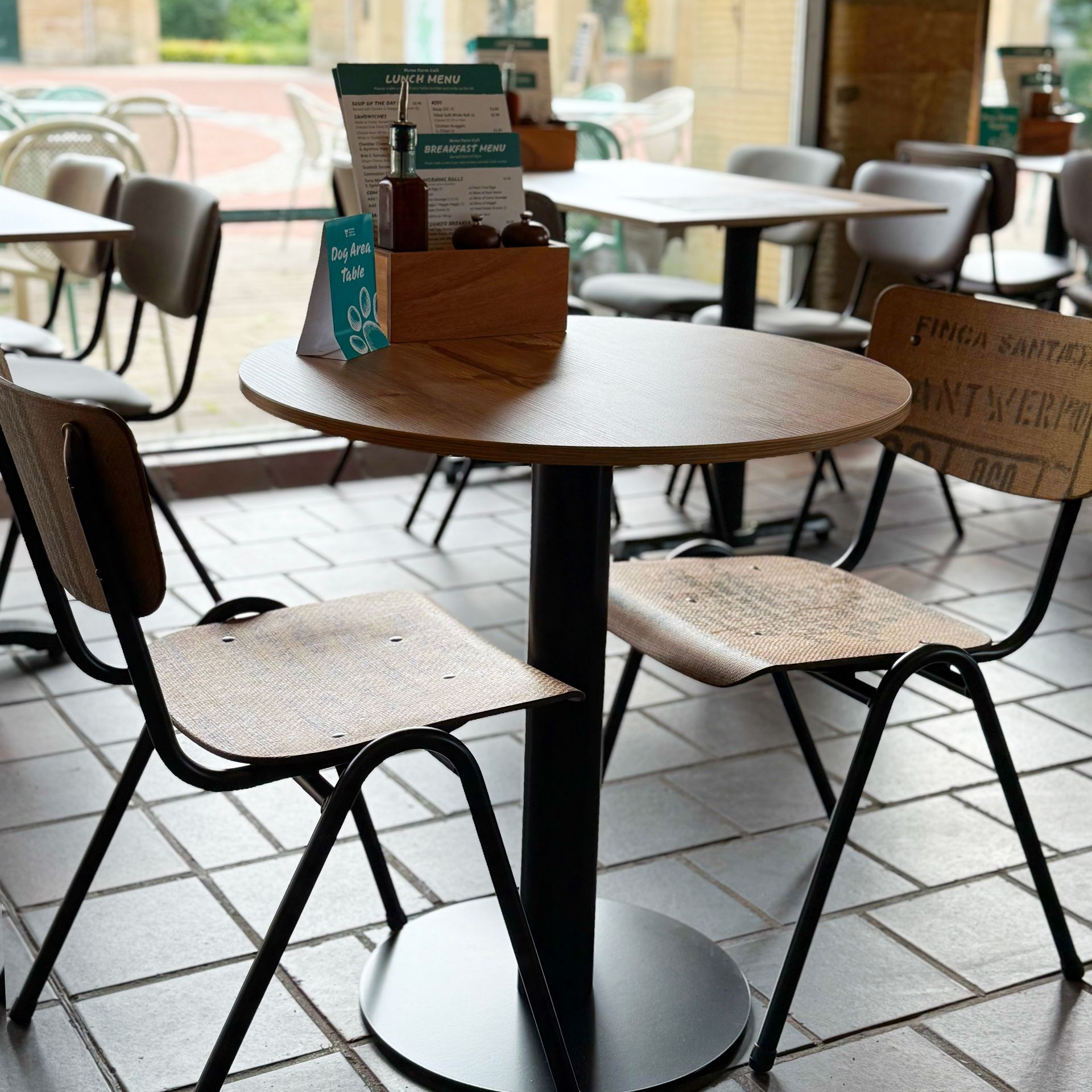Cafe table and chairs near a window. Wooden tables, black metal frames, and tiled floor.