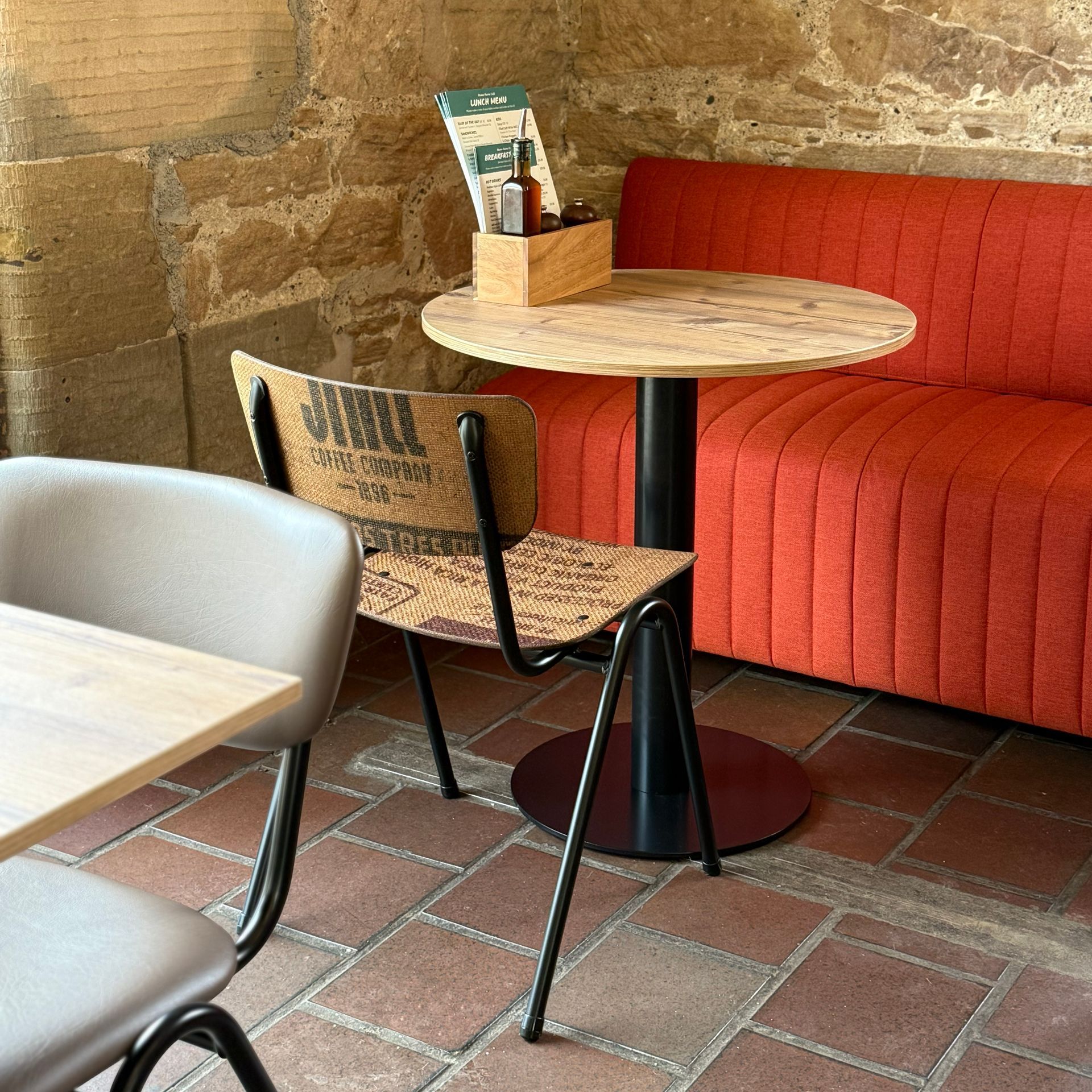 Restaurant seating: round table, orange booth, chair with coffee sack back, brick floor, and stone wall.