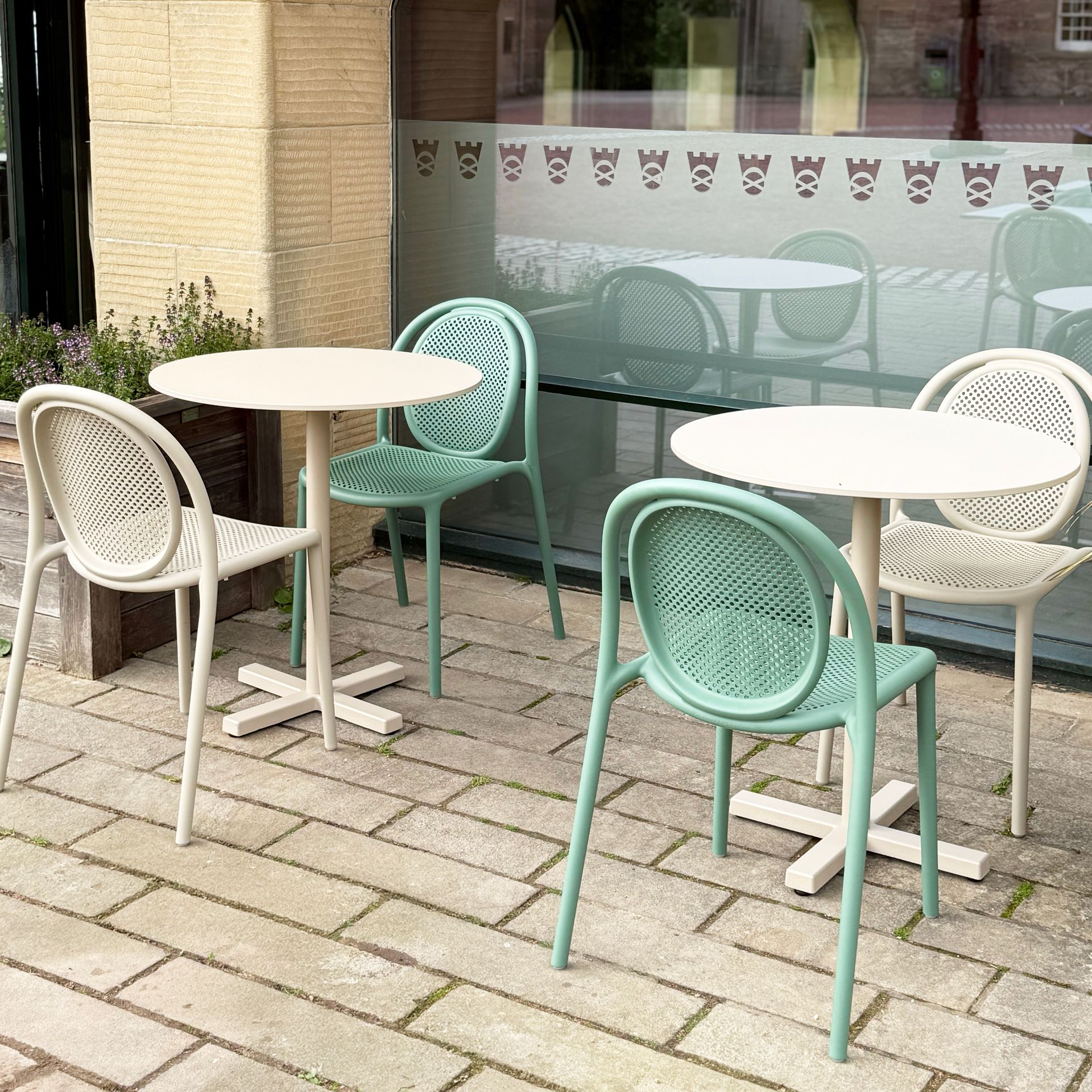 Outdoor cafe seating: round tables, woven-backed chairs in green and white on a brick patio.