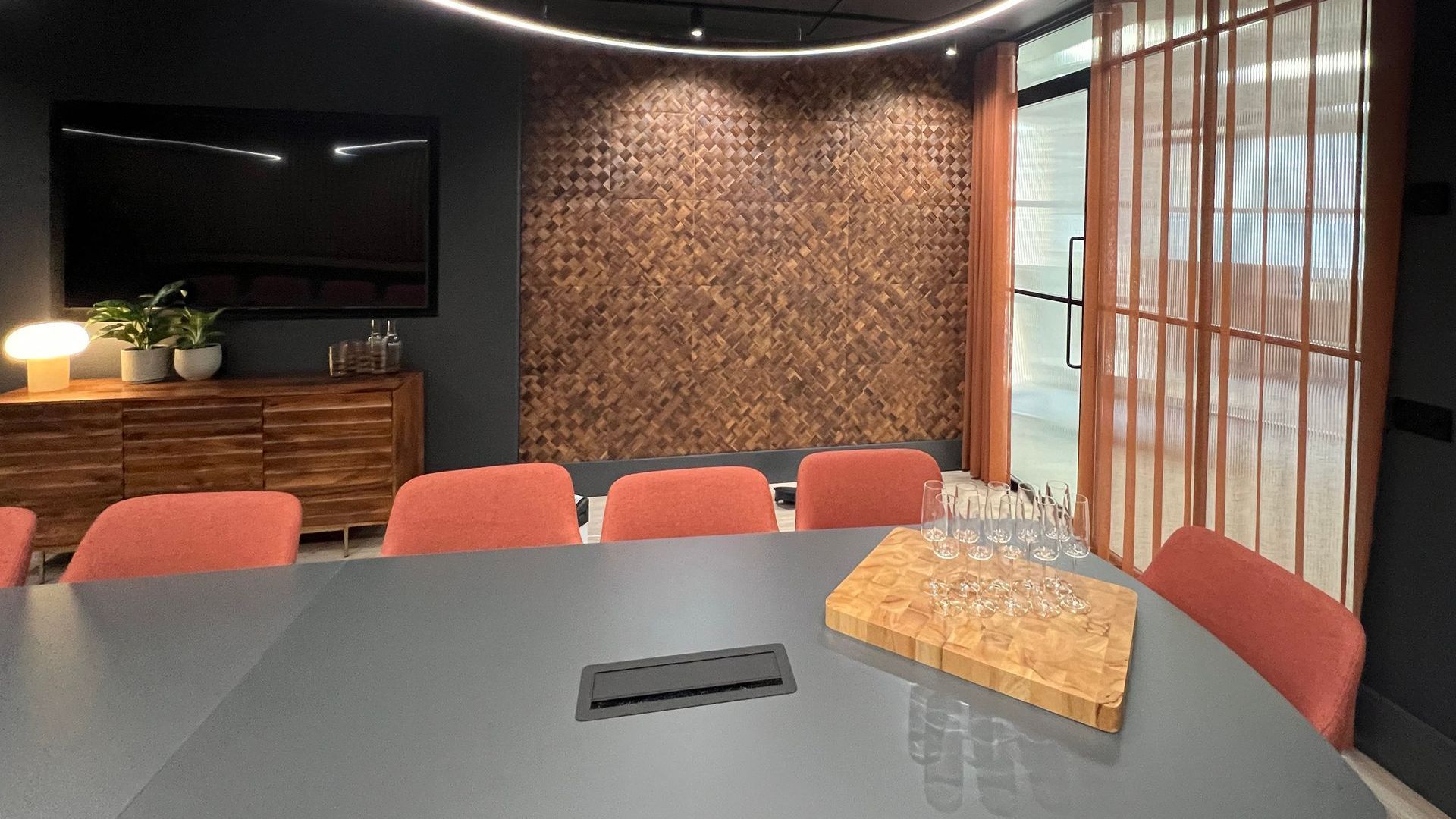 an innovative boardroom with a large grey board table, warm wooden storage, orange soft seating, and sustainably manufactured wall coverings made from banana tree bark.