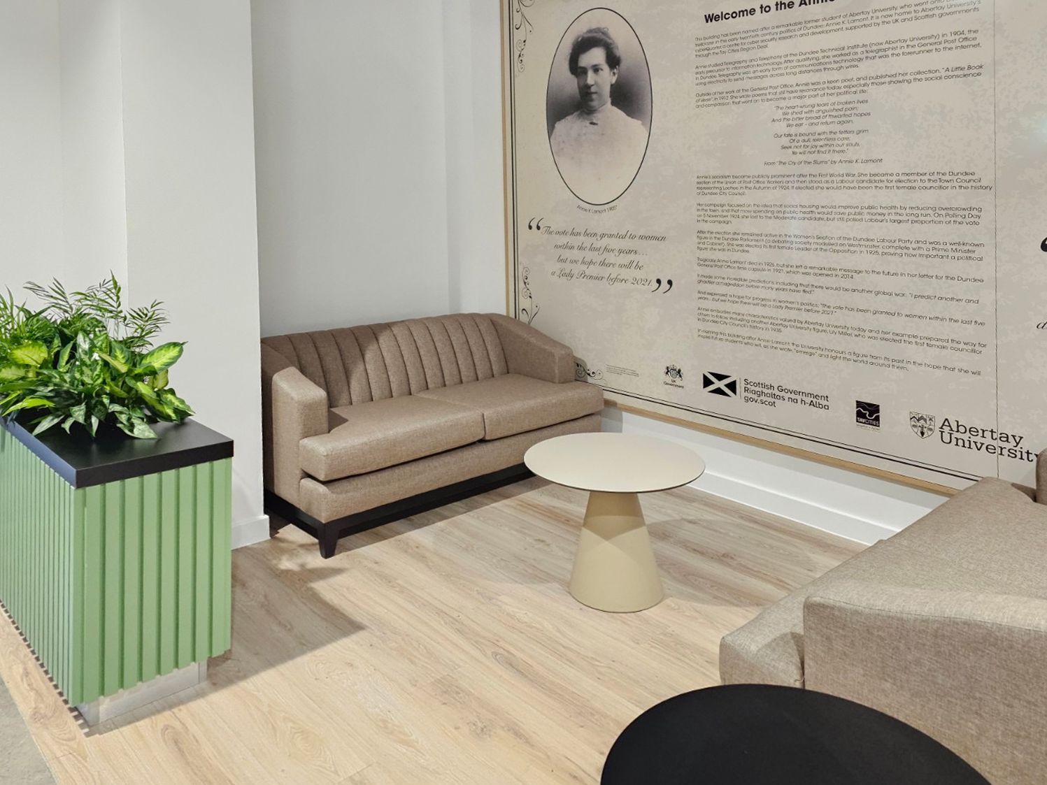 Corner of the cafe with beige sofas, a small circular table, a plant divider, and a mural with information about Abertay University. 