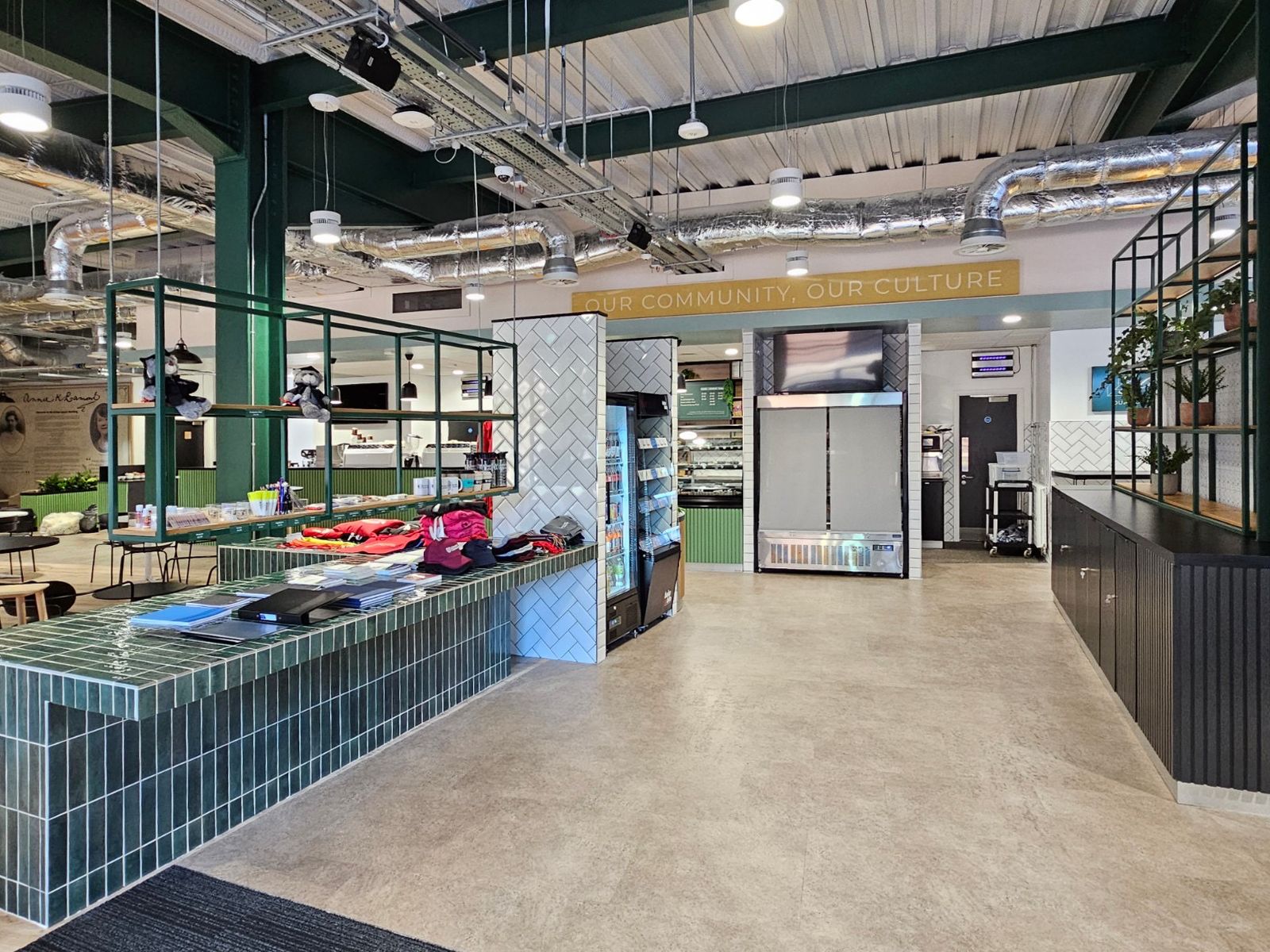 Space for people to get a drink, food, and snacks from the fridge, with a green tiled counter to look at merchandise. On the other side of the space is a black dividing unit with storage cabinets and shelving for plants. 