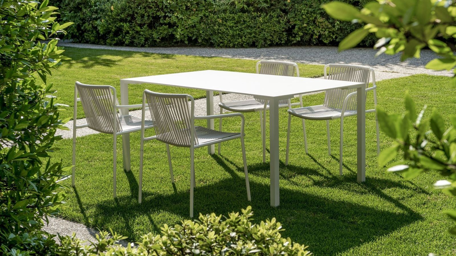 a white table sits outside on top of healthy green grass. there are four white chairs at the table. in the background, there are healthy bushes and trees. 