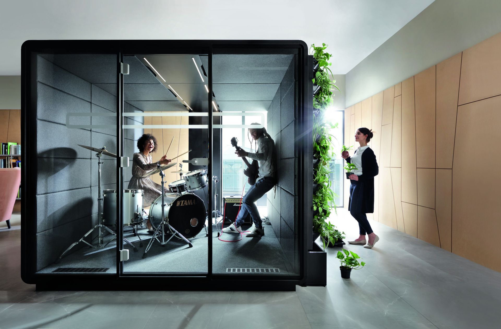 Band playing inside soundproof booth with a person observing outside. Modern office setting, light colors.