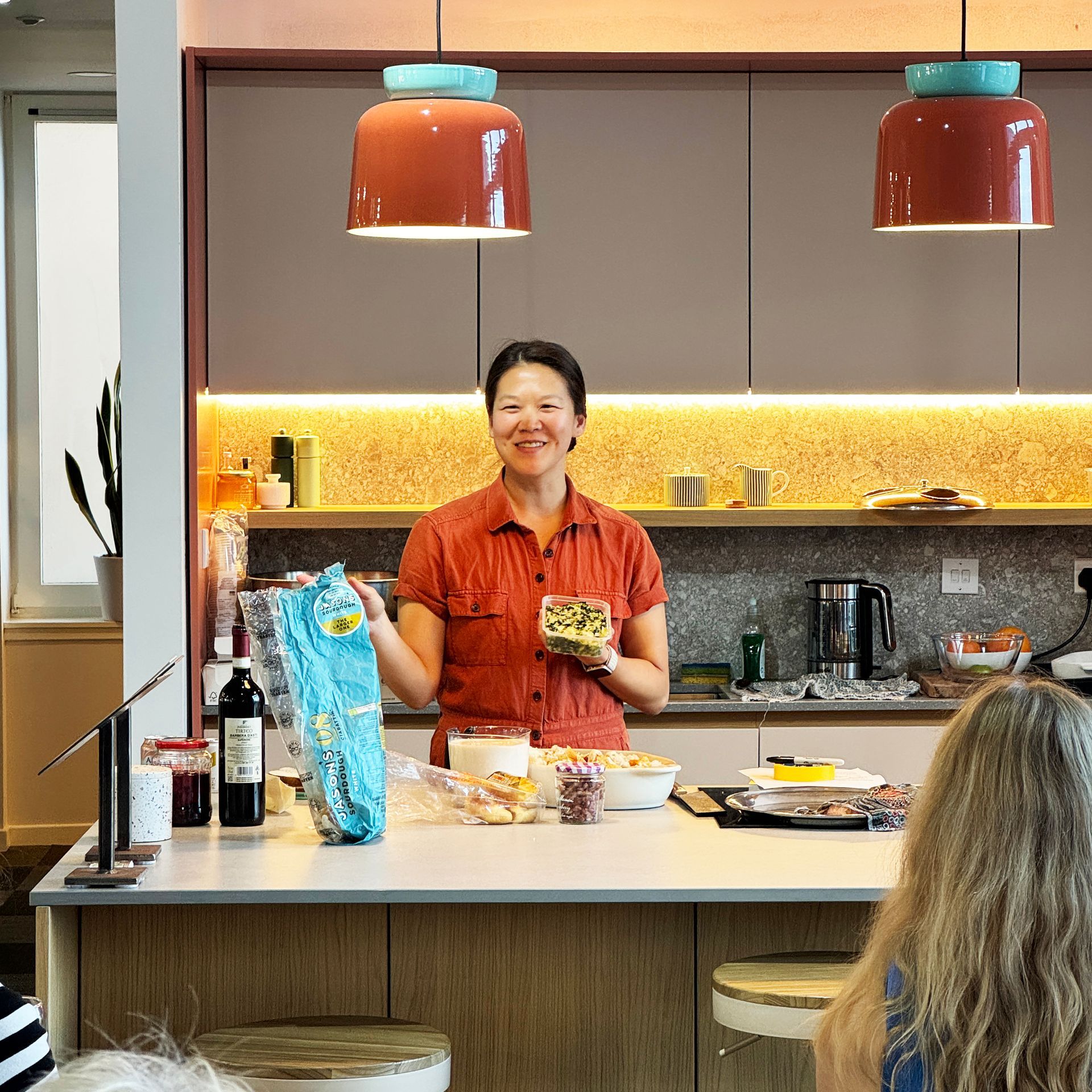 Woman in kitchen holding food, smiling. Two copper pendant lights, counter with ingredients, audience.