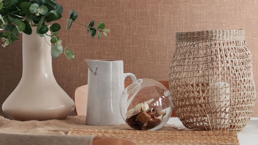 Peach toned woven-style wallpaper sits behind a table. On top of the table are vases and a basket with a candle inside. 