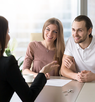 Woman in black jacket gestures to smiling couple at a table; keys and documents present.