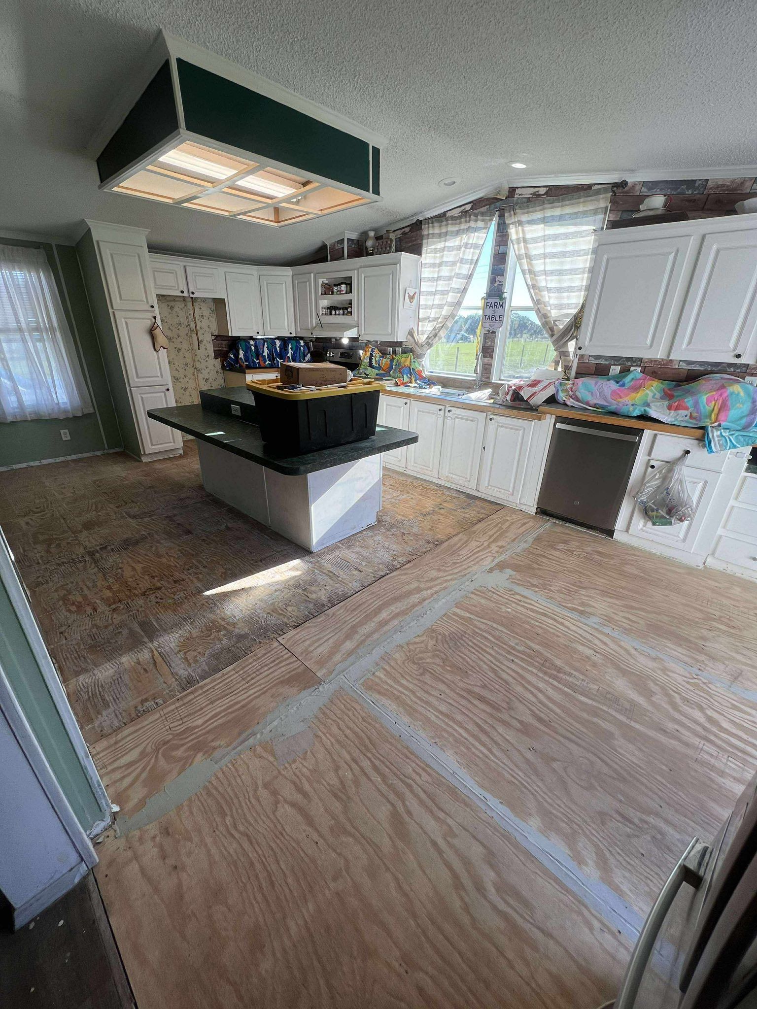Kitchen with plywood flooring, white cabinets, island, and sunlight streaming through windows.