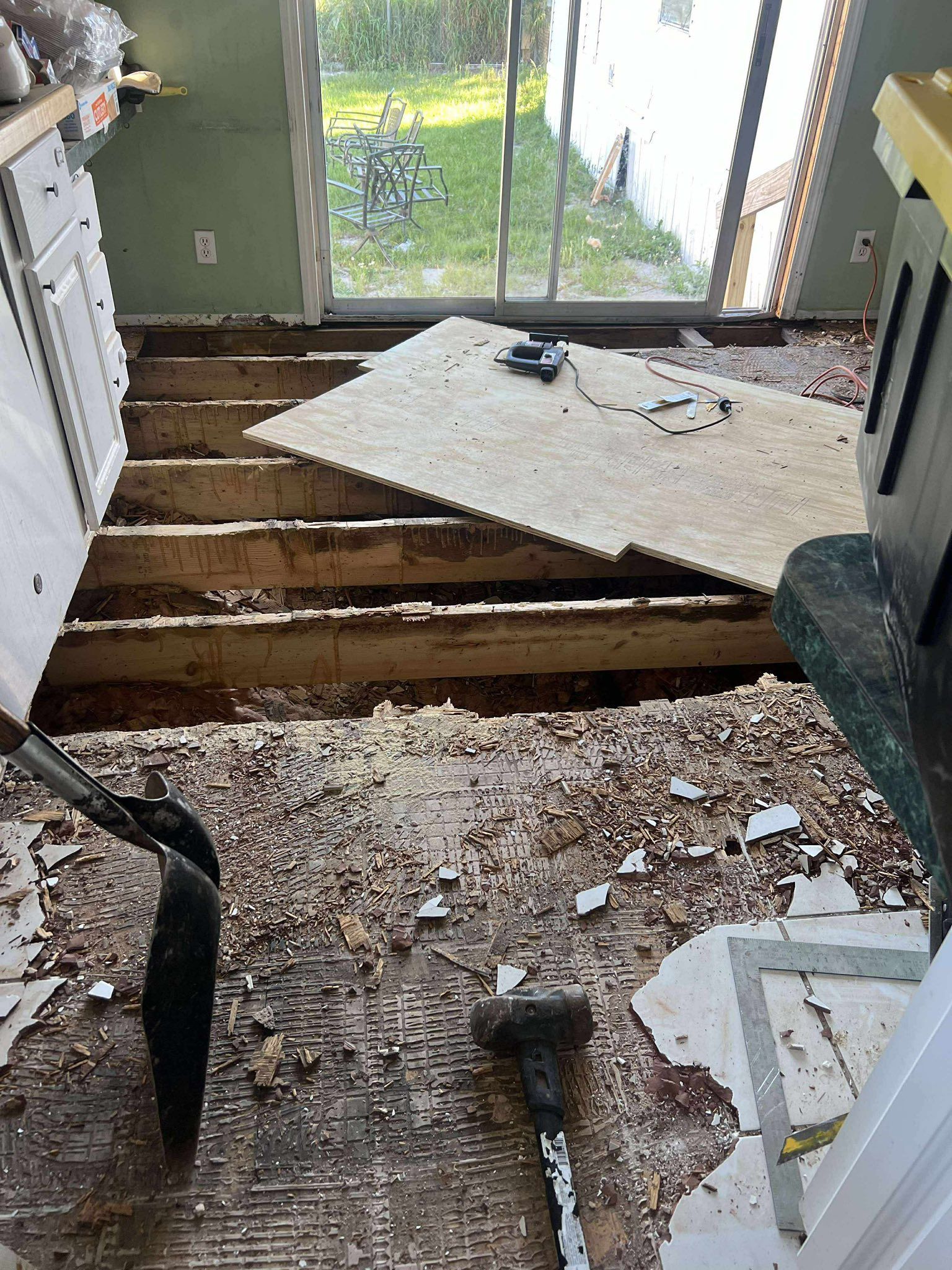 Kitchen floor in disrepair with exposed beams, demolition tools, and removed flooring near sliding glass door.