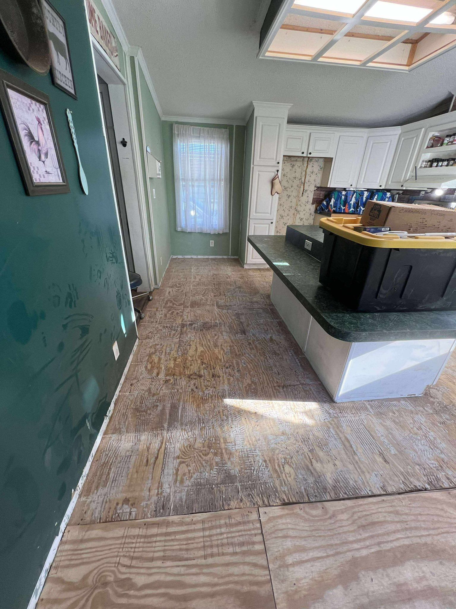 Interior of a kitchen with damaged flooring. Green wall on the left, cabinets on the right, and a window in the distance.