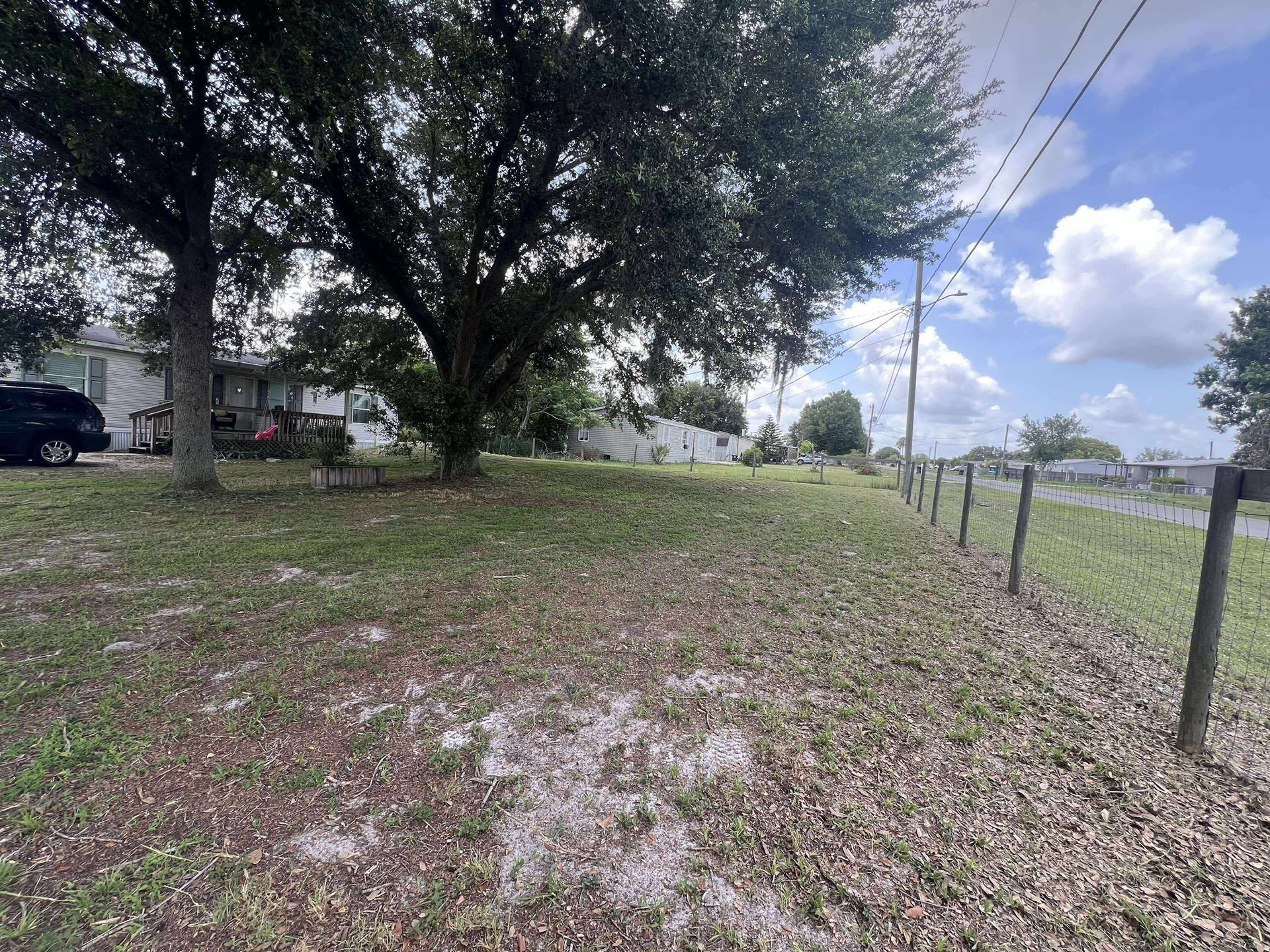 Grassy yard with fence and a house under a tree, blue sky with clouds in the background.
