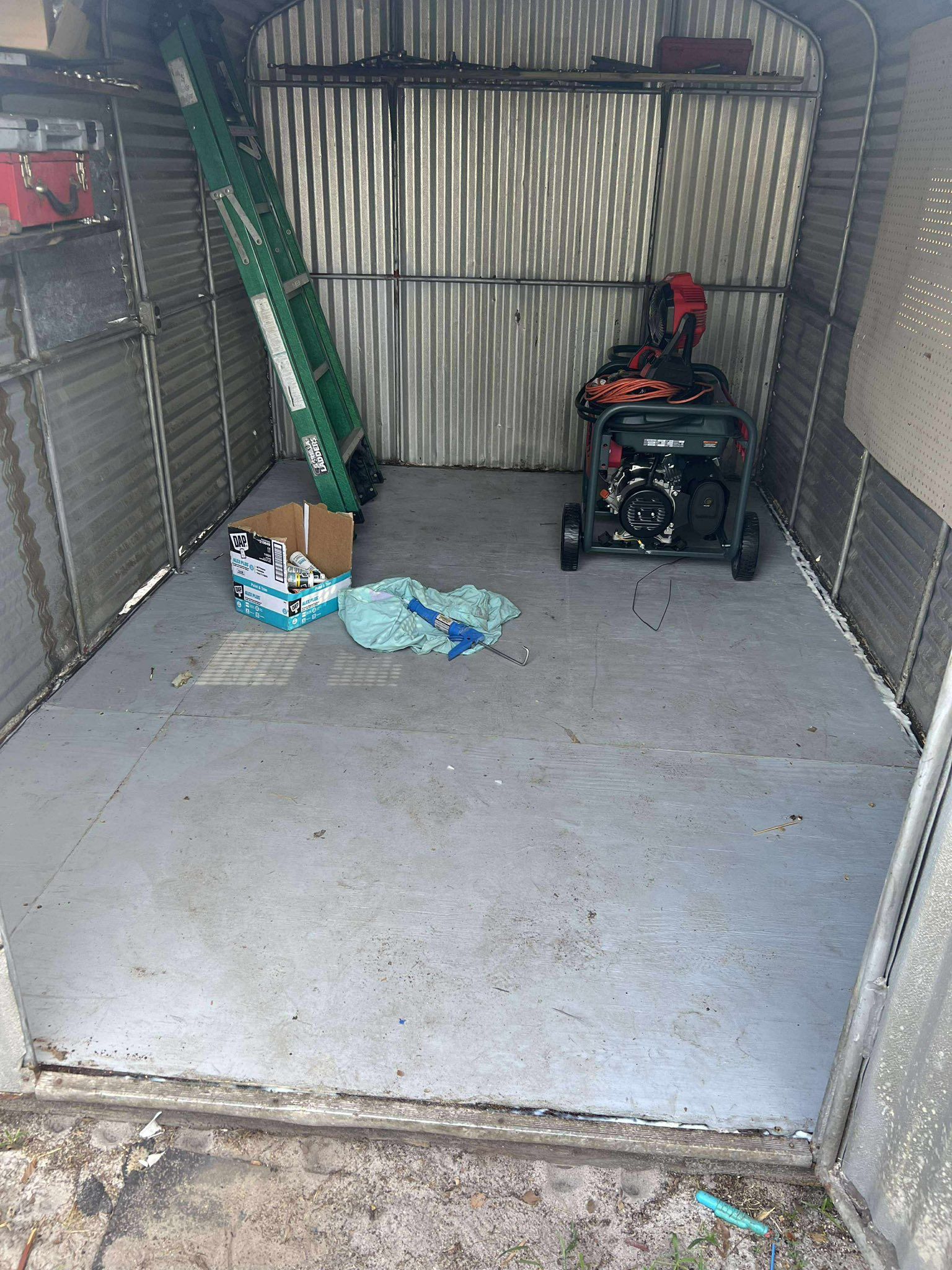 Inside a metal shed: concrete floor, green ladder, generator, cardboard box, and a blue cloth.