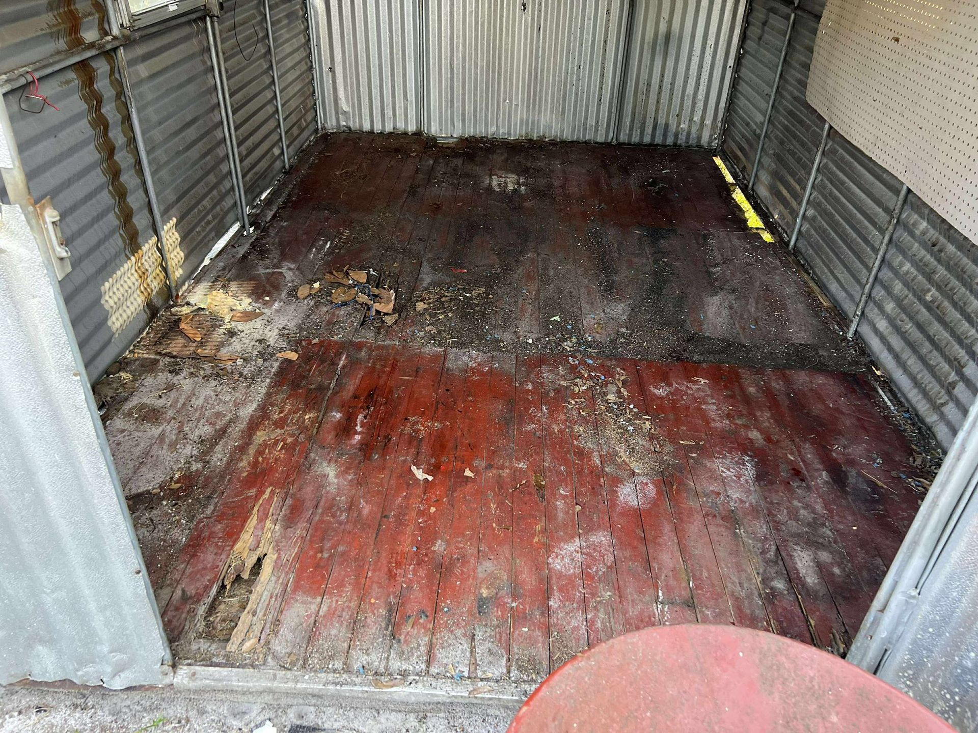 Interior of a dilapidated shed with a dirty red wood floor and corrugated metal walls.
