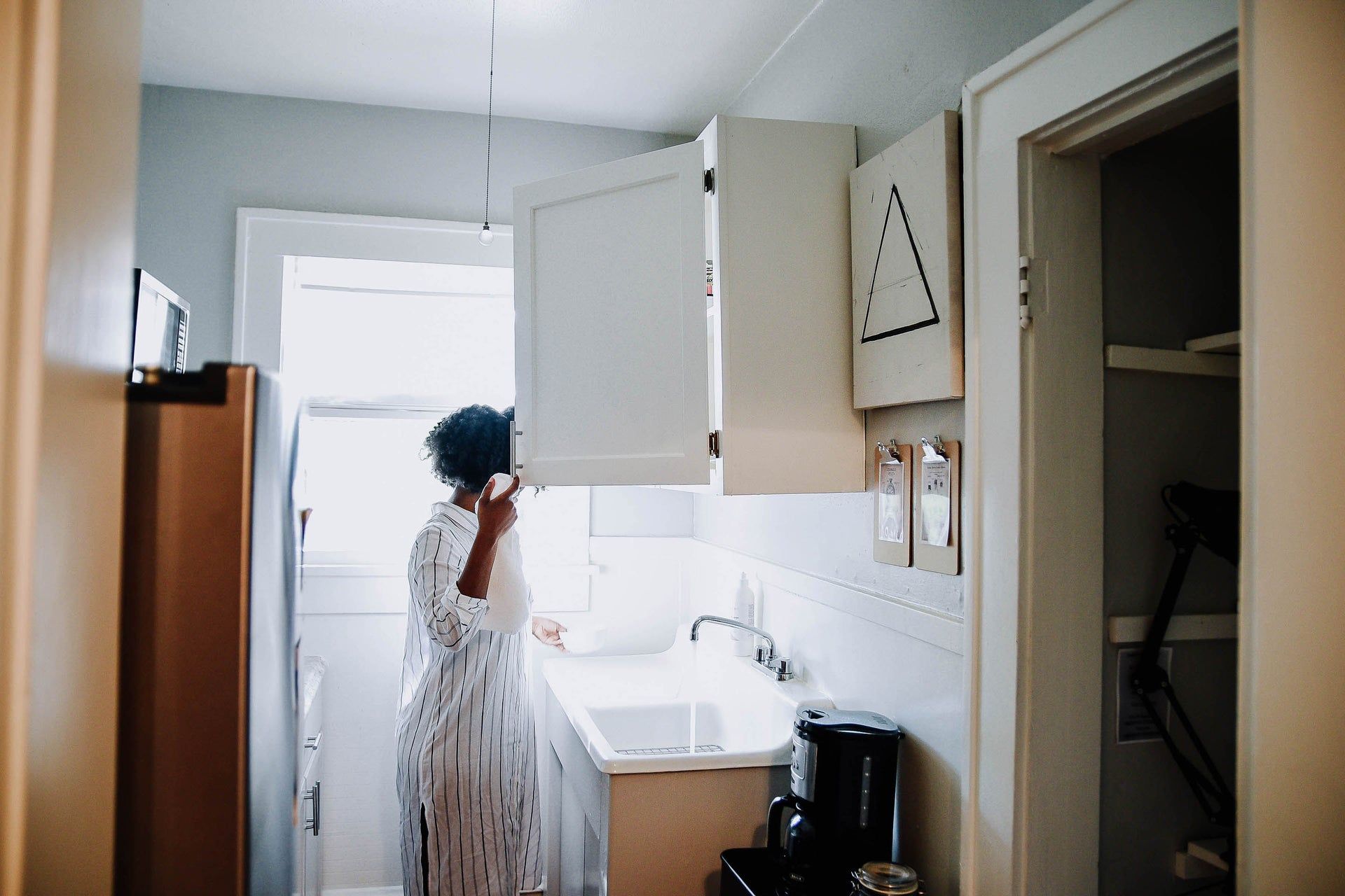 Person in white dress standing at a kitchen sink in a bright, open cabinet kitchen