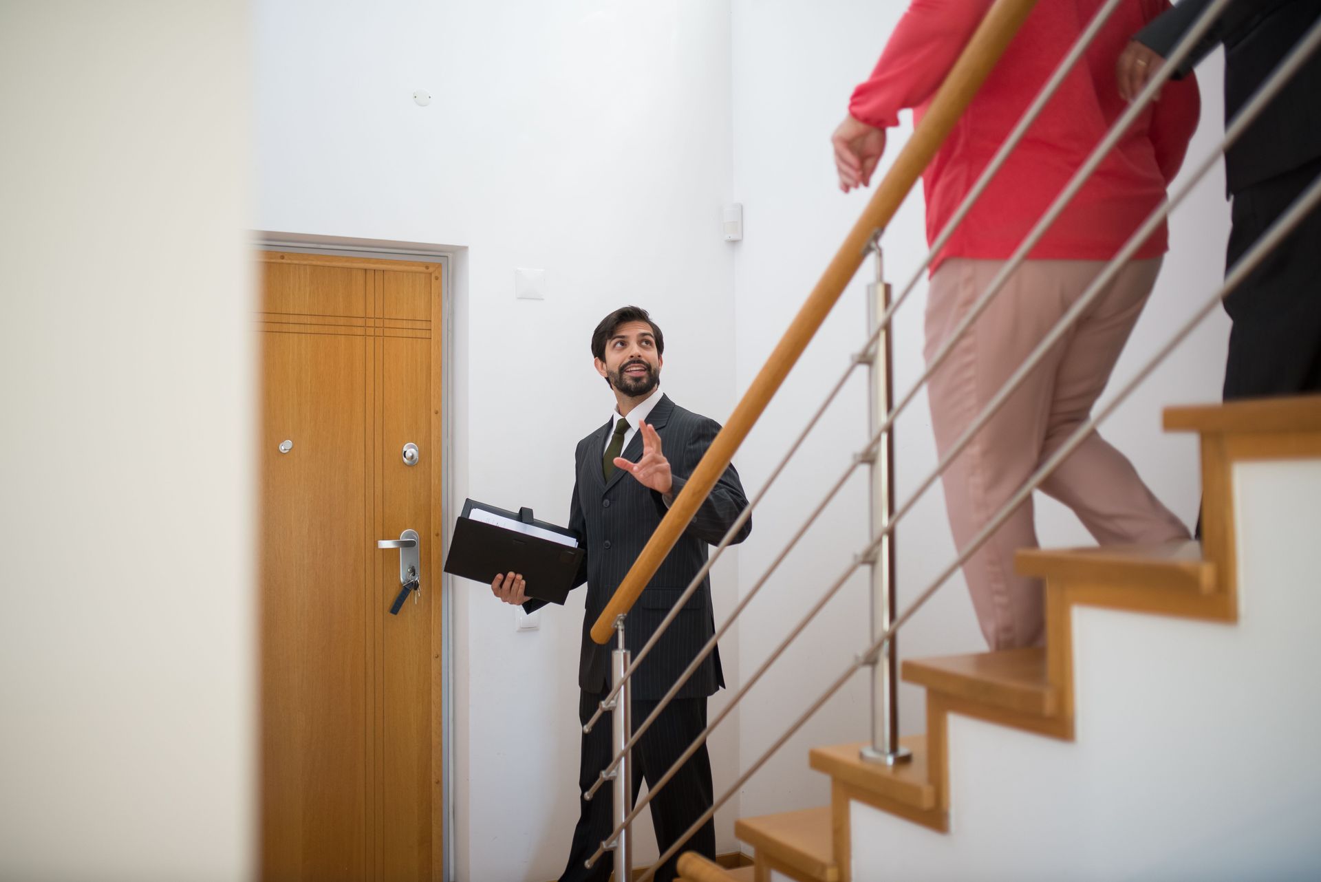 Man in a dark suit carries a box near a wooden staircase as another person in a red shirt descends.