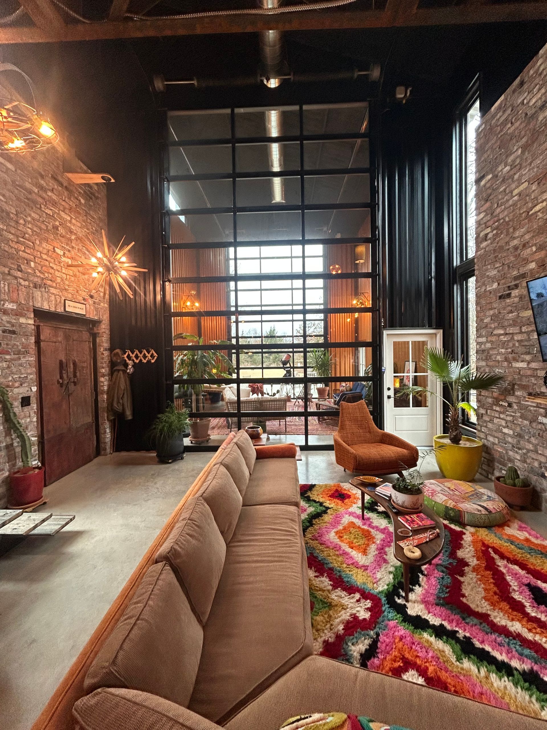 A living room with stone walls, a long brown sofa, a colorful patterned rug, and a tall industrial garage-style window.