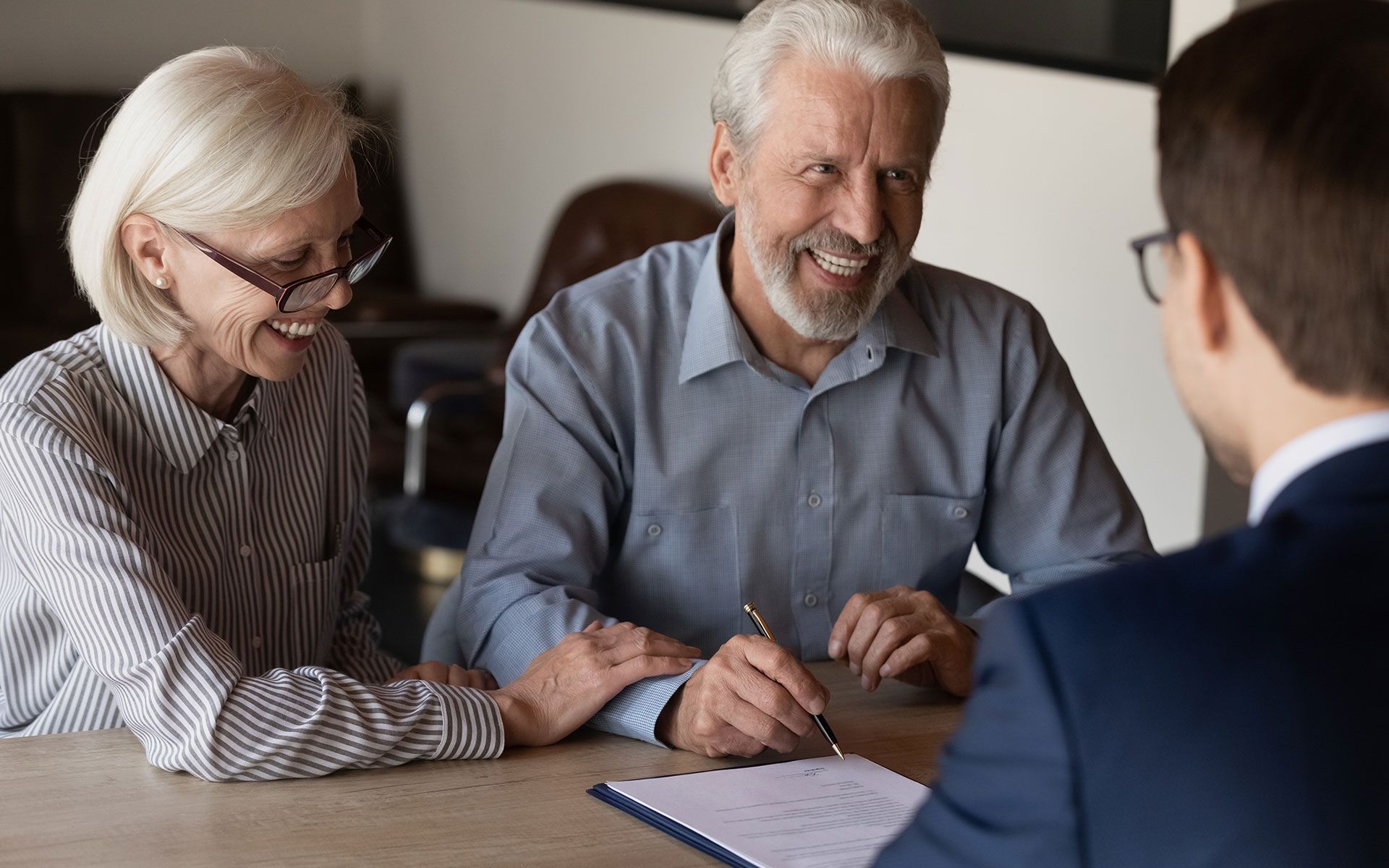 An elderly couple is sitting at a table signing a document with a man.