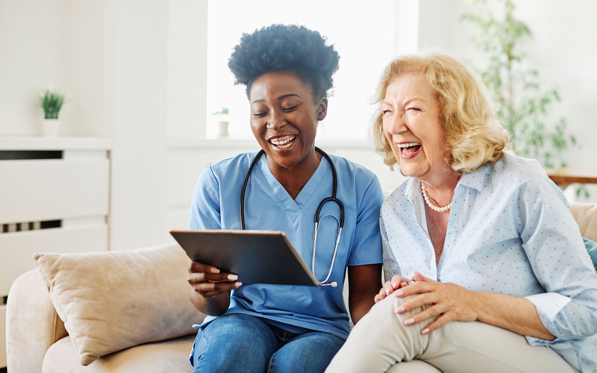 A nurse is sitting next to an elderly woman on a couch looking at a tablet.