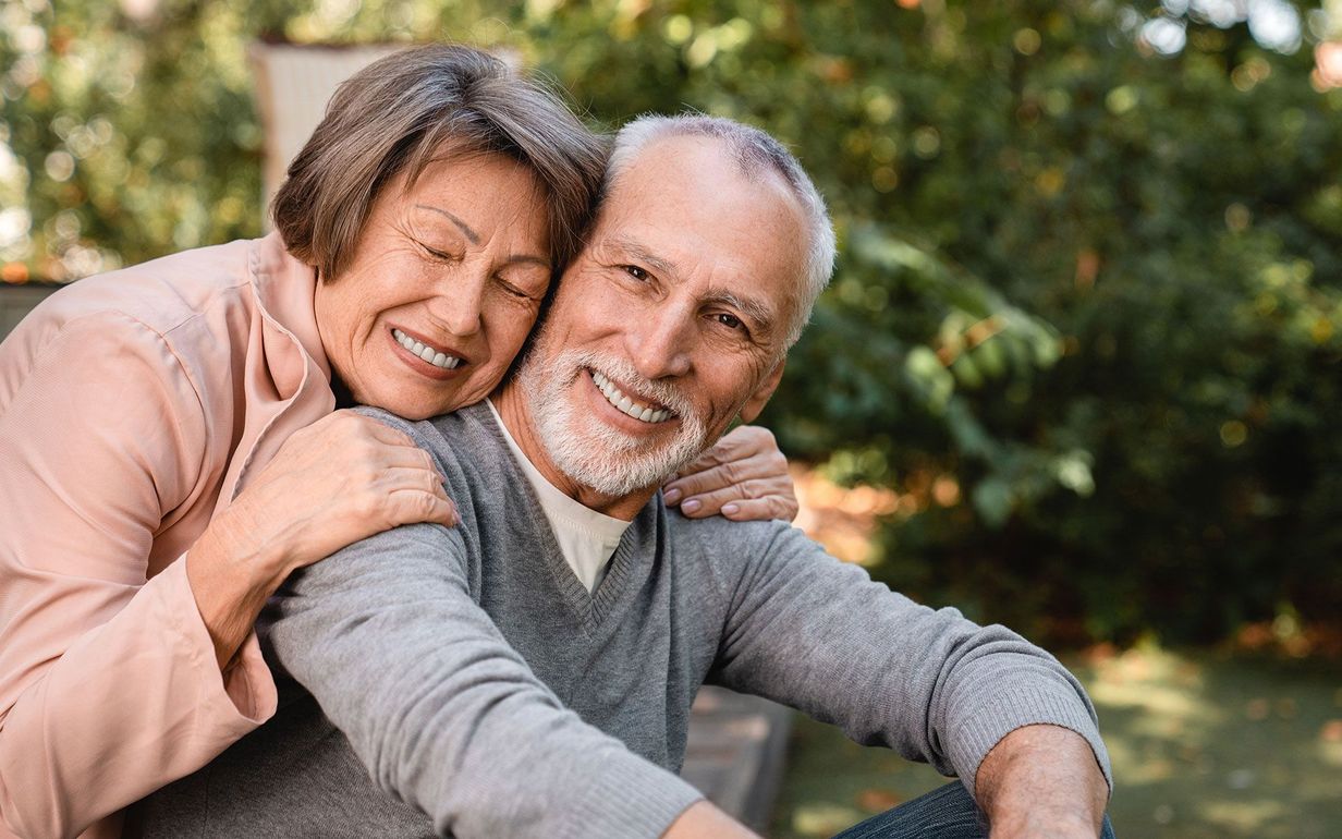 An elderly couple is hugging each other and smiling while sitting on a bench.
