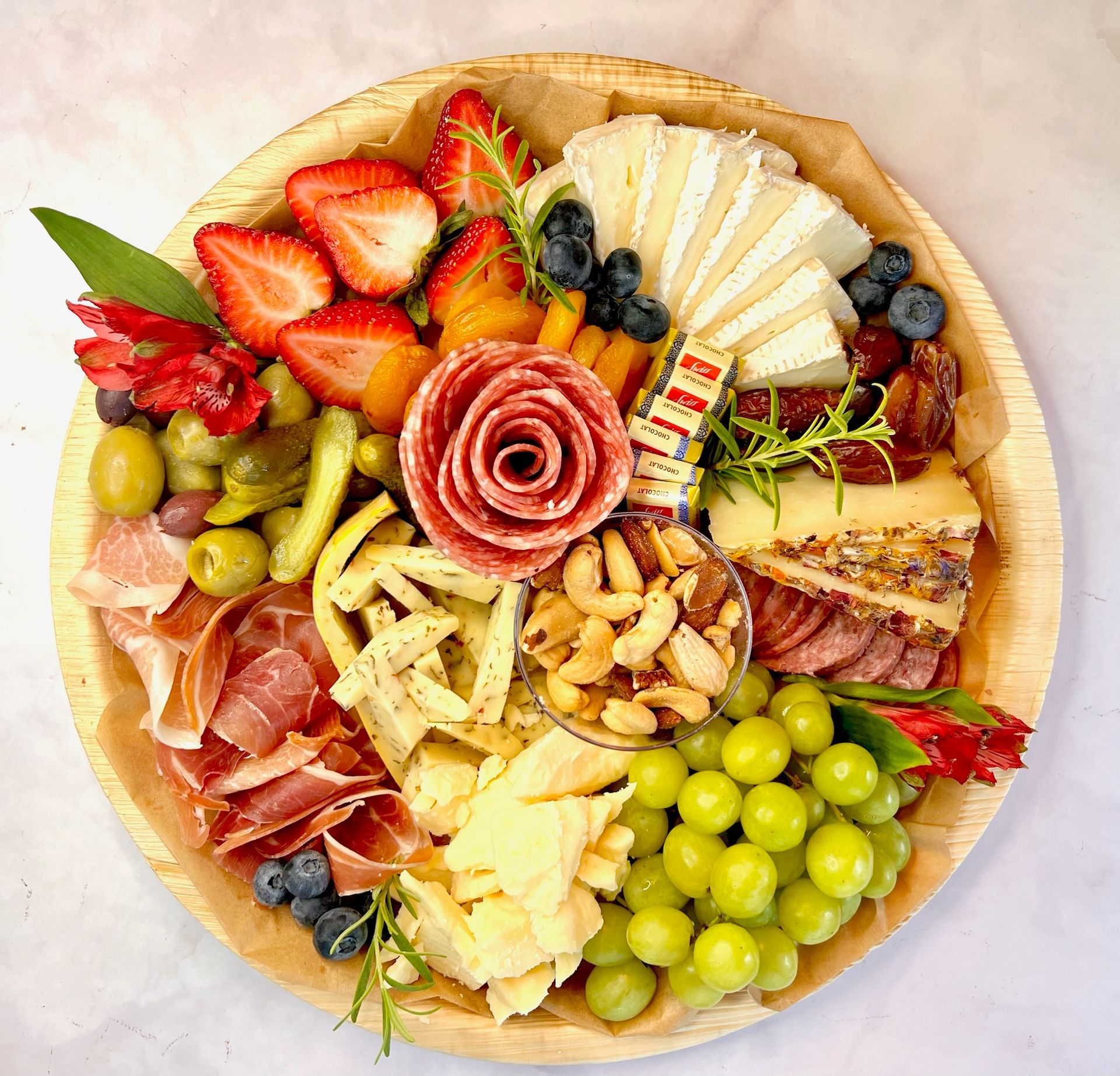 A wooden plate topped with meat , cheese , nuts , grapes and strawberries.