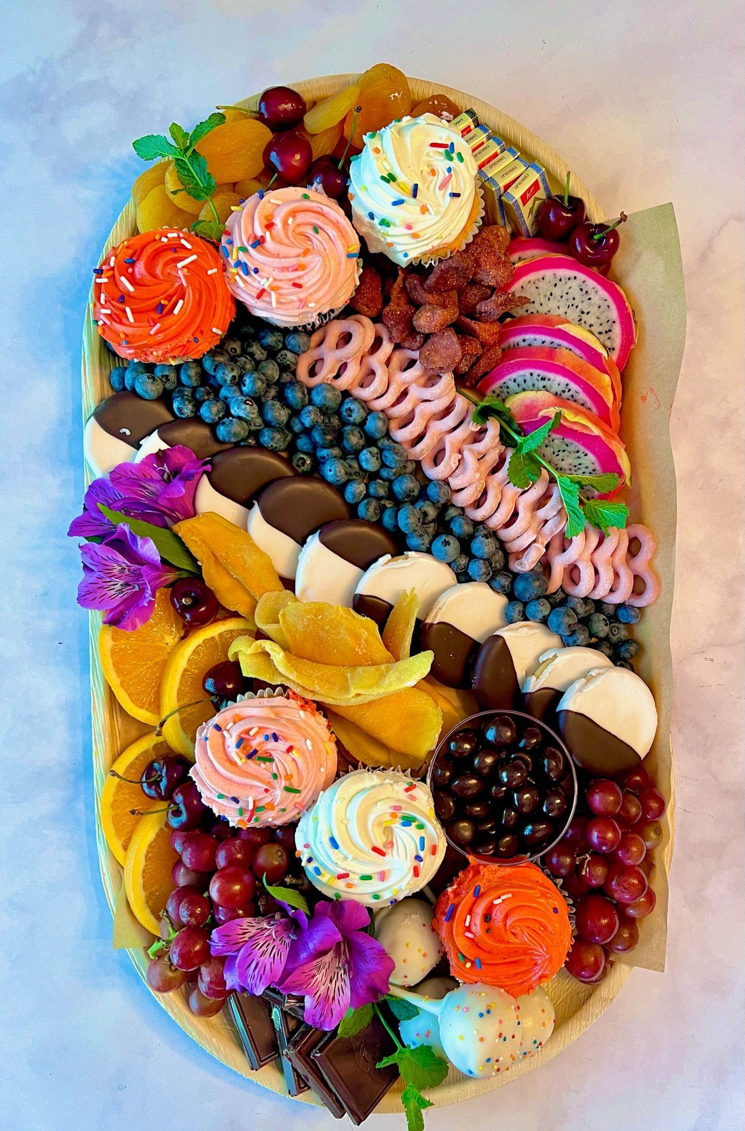 A wooden tray filled with fruits , cupcakes , and flowers.