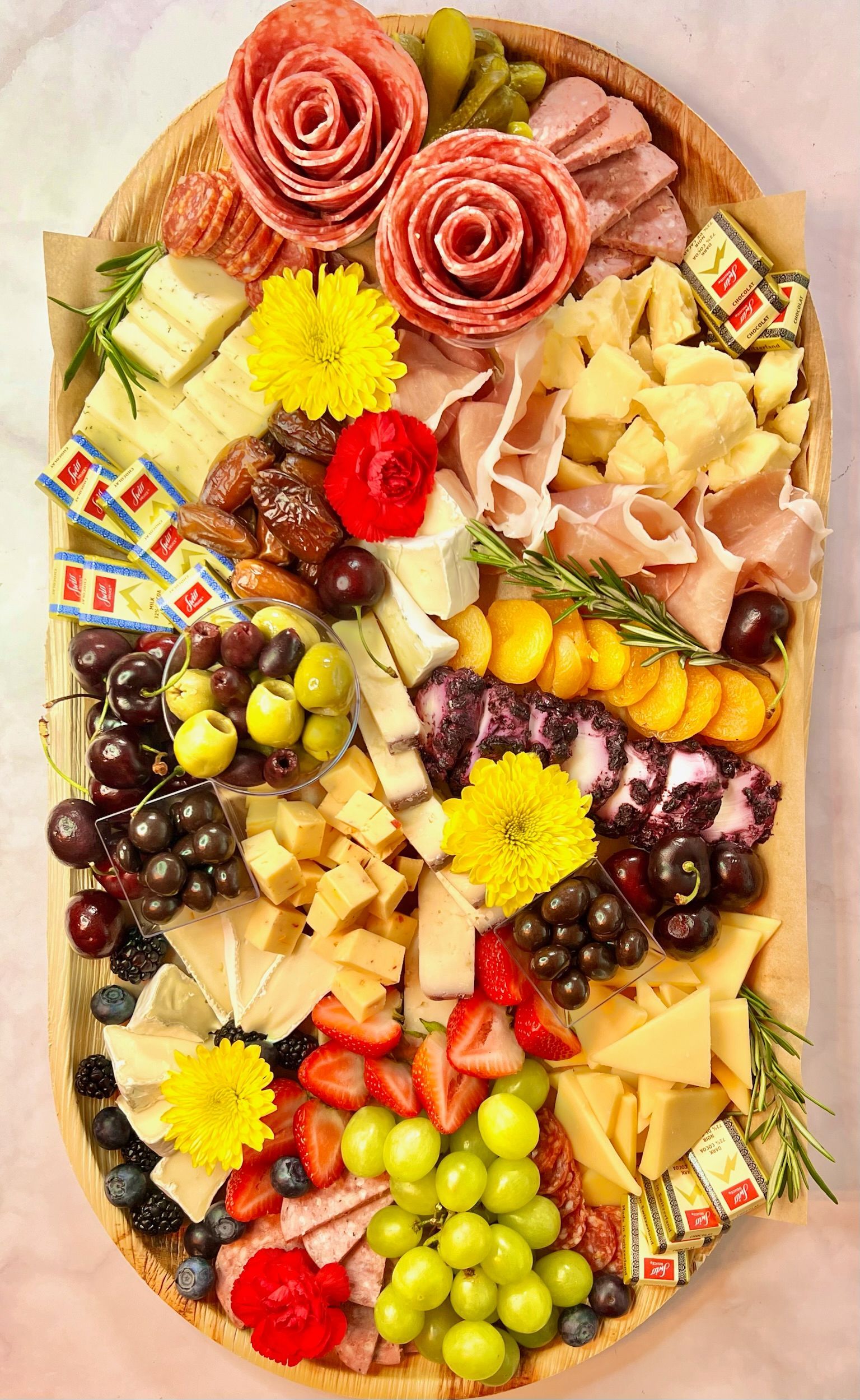 A wooden tray filled with meats , cheeses , grapes , and flowers.