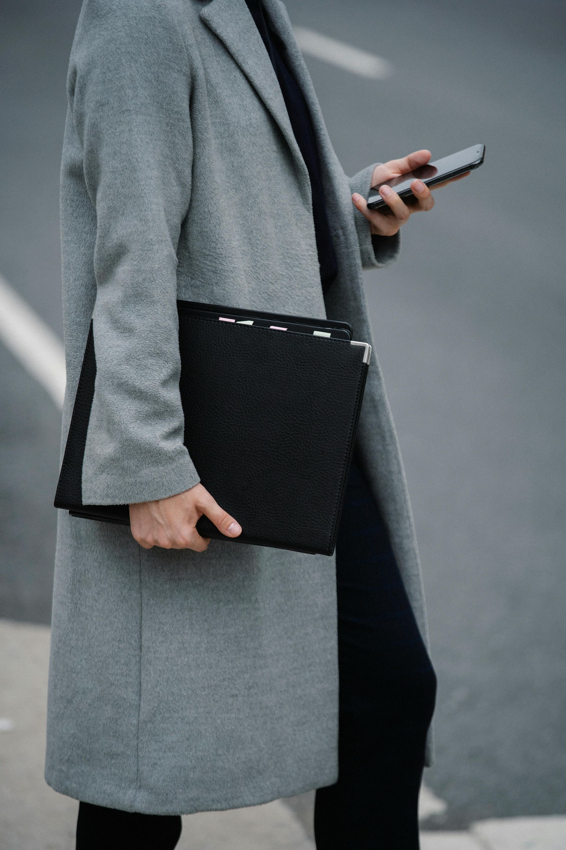 Person in grey coat holding a black folder and a phone while walking on a street.