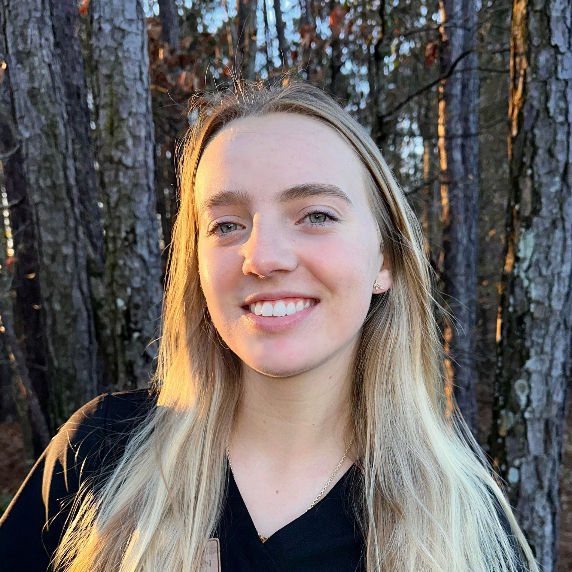 A young person with long blonde hair smiles outdoors in front of a forest during sunset, with light hitting their face.