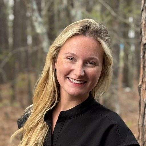A person with long blonde hair and a friendly smile, wearing a black shirt, posing in a blurred outdoor forest setting.