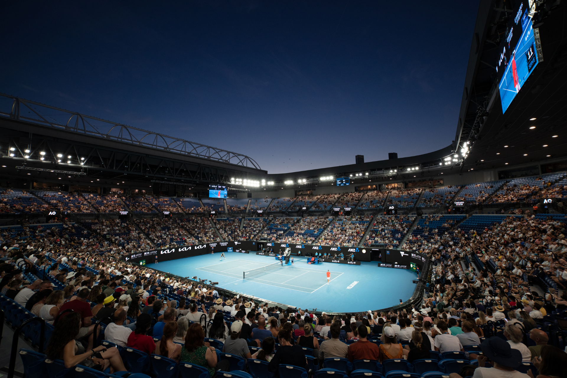A stadium filled with people watching a tennis match at night.