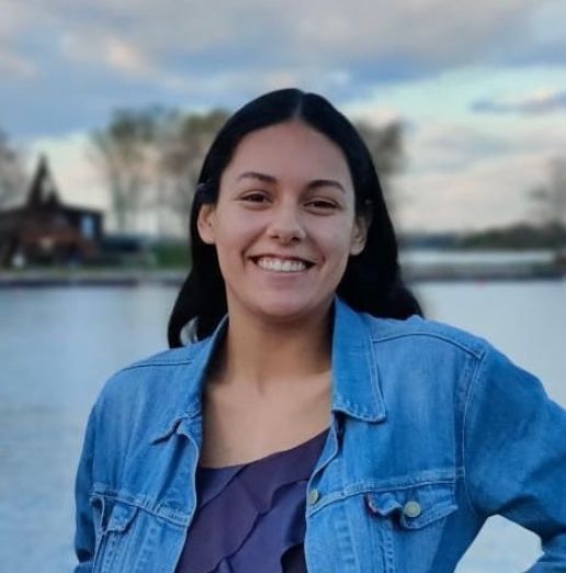 A woman wearing a denim jacket and a purple top is smiling in front of a body of water.